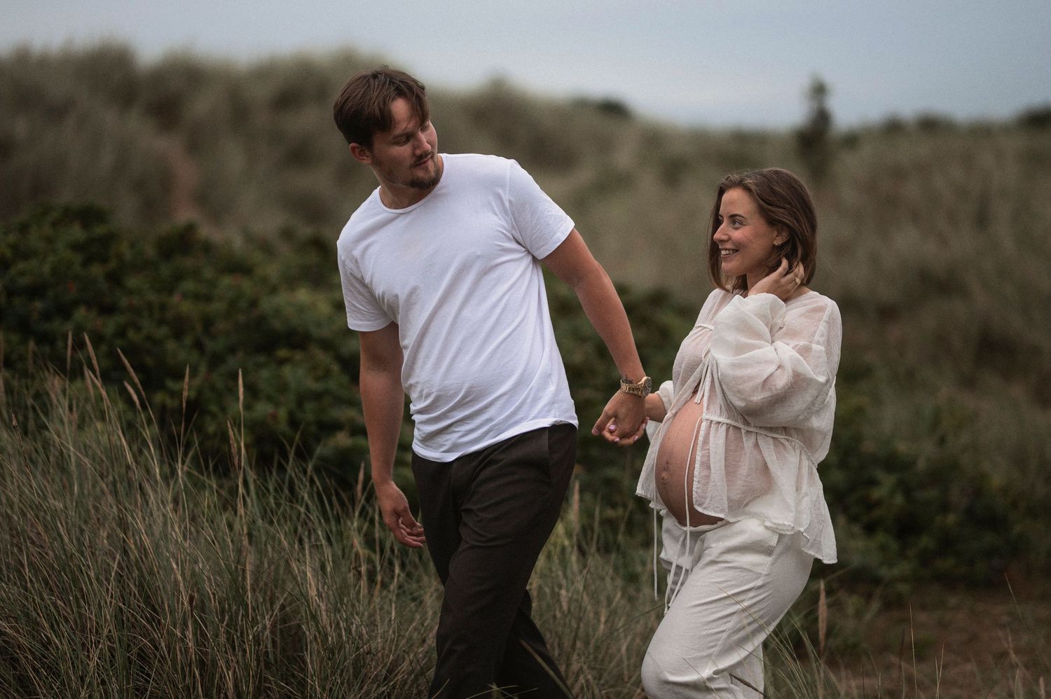 A couple walks hand in hand along a coastal path during their maternity shoot.