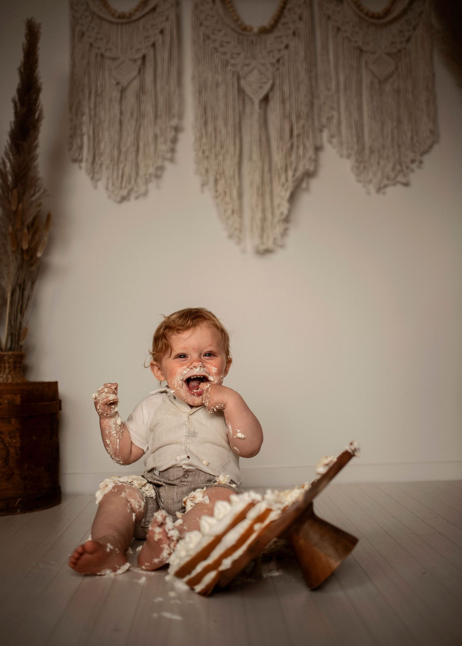 A messy cake smash photoshoot showing a baby covered in frosting while sitting on a wooden floor with macrame decor.