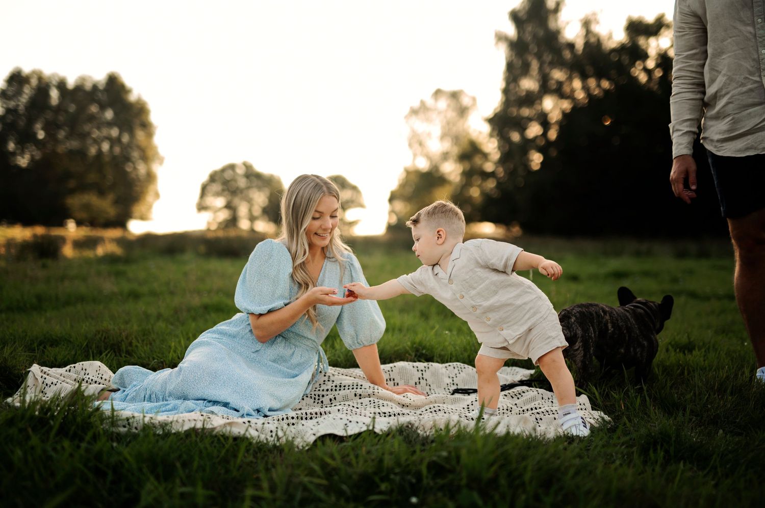 Playful interaction on picnic blanket with black dog in background during golden hour.