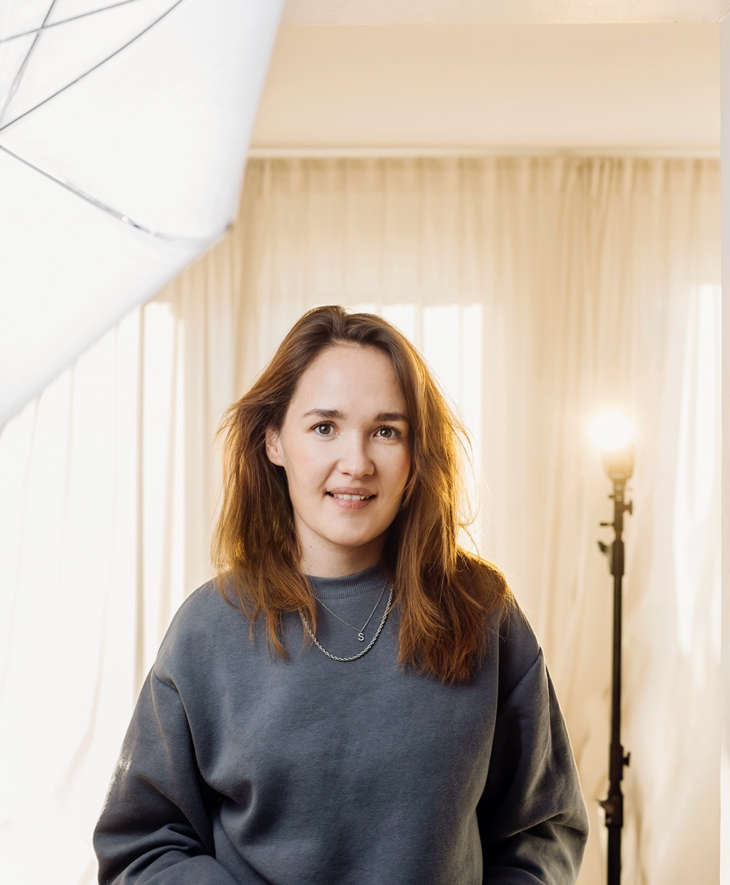 Young woman with auburn hair smiling in photography studio wearing gray sweatshirt with lighting equipment.