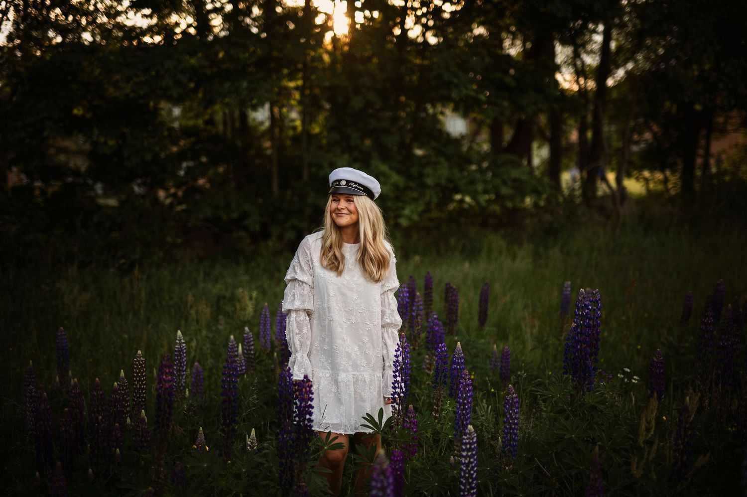 A young woman in a white dress and sailor cap stands among purple lupine flowers at sunset.