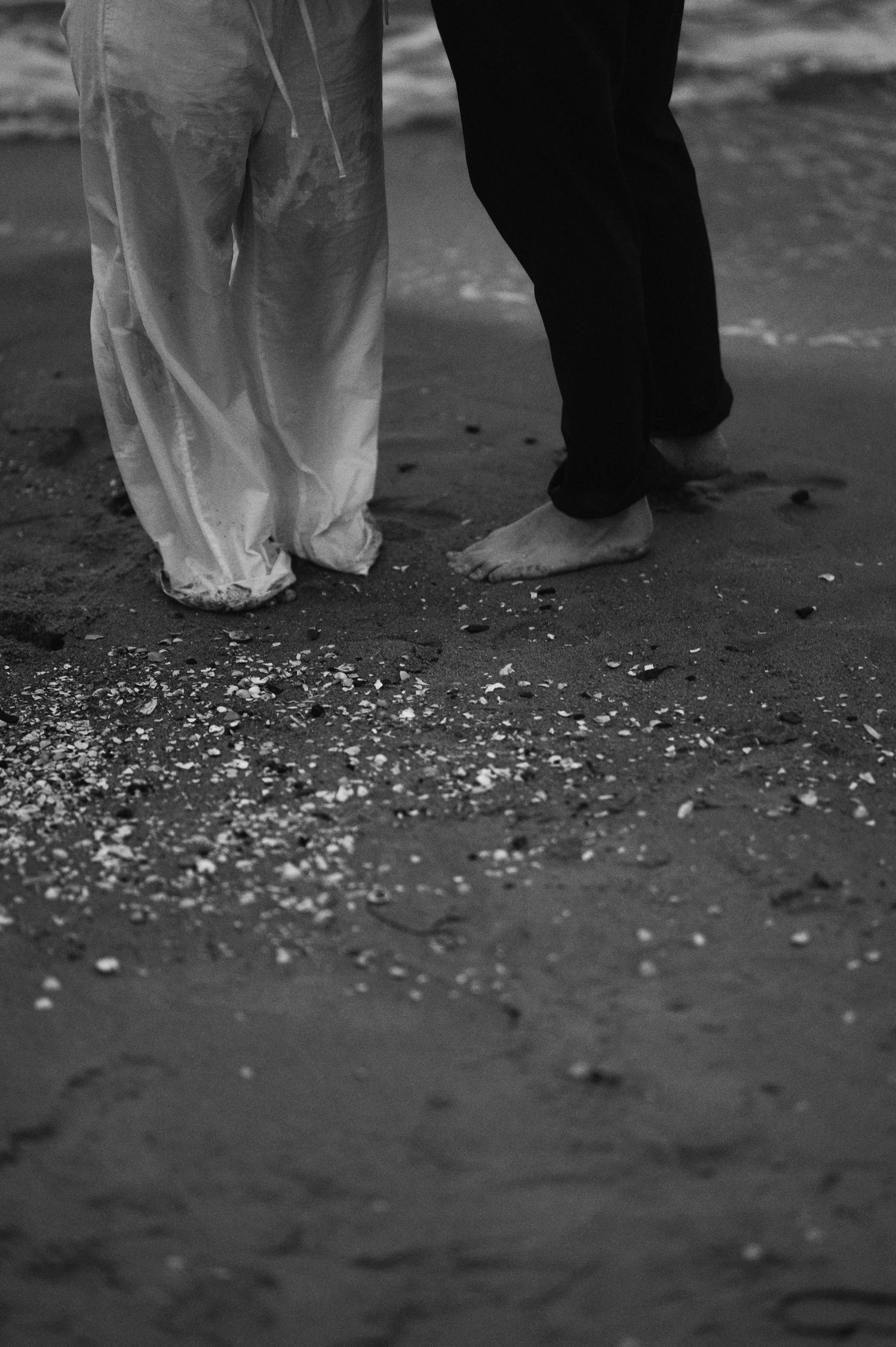 Black and white photo of feet standing in wet sand with ocean waves washing ashore.
