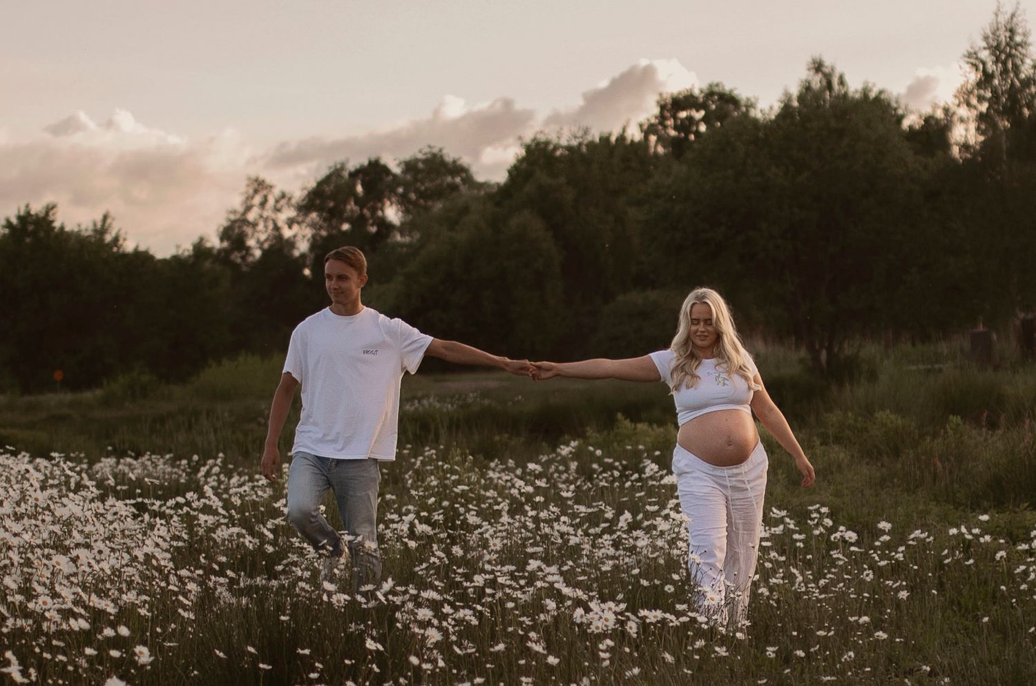 A couple walks through a blooming white field of wildflowers during golden hour at sunset in the countryside.