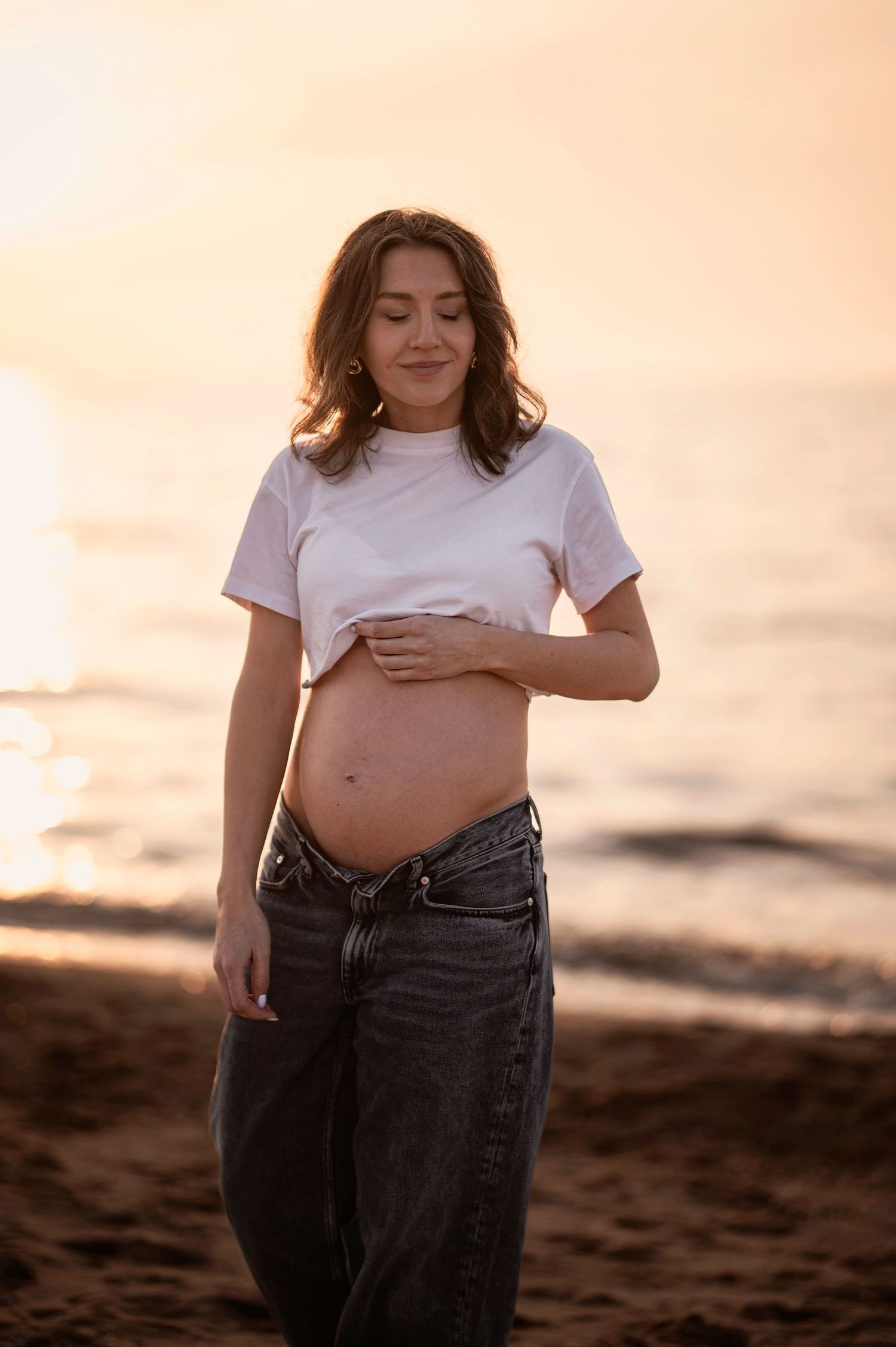 Expectant mother in white t-shirt and jeans poses on beach during golden hour sunset.
