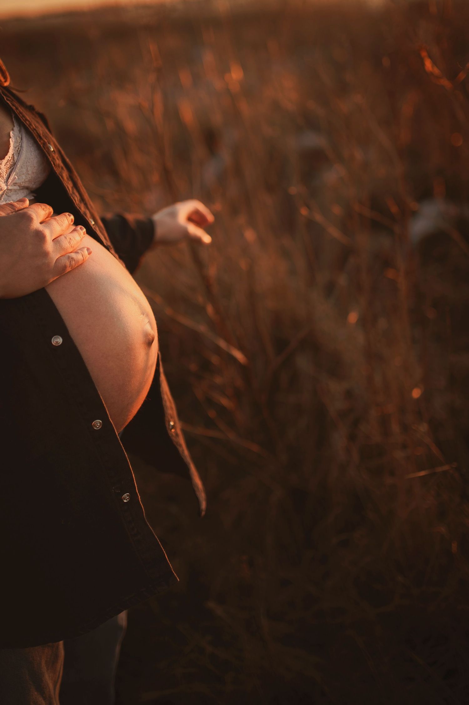 Close-up of a pregnant belly in dark clothing against autumn grass at golden hour.