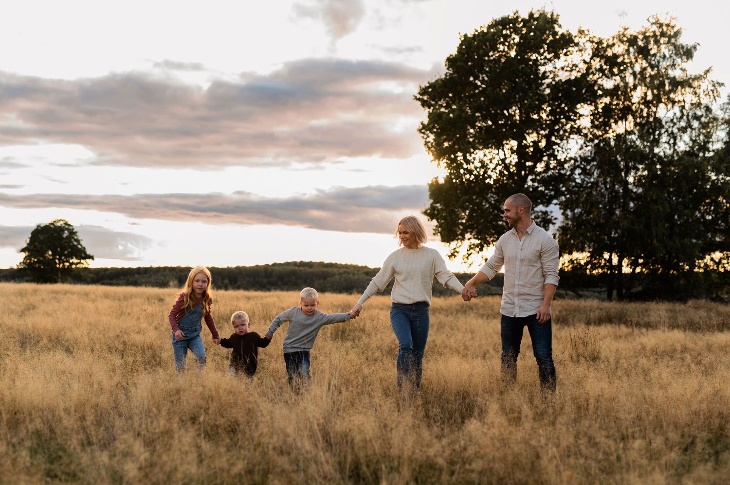 Family holding hands while walking through golden field at sunset.