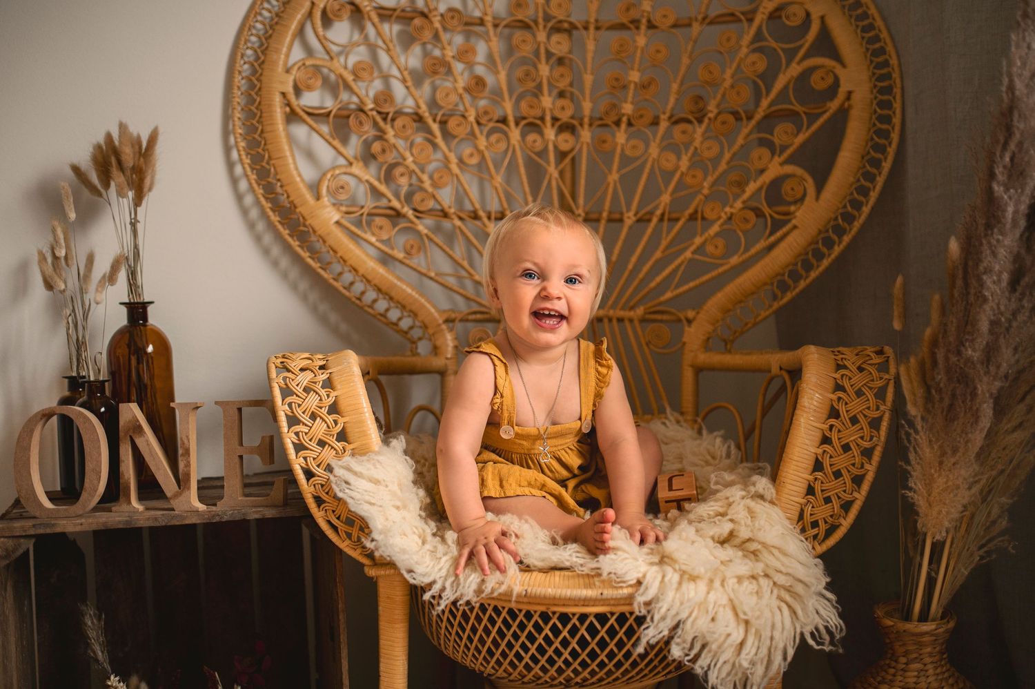 Baby wearing yellow outfit smiles while sitting in vintage peacock chair with boho decorations.