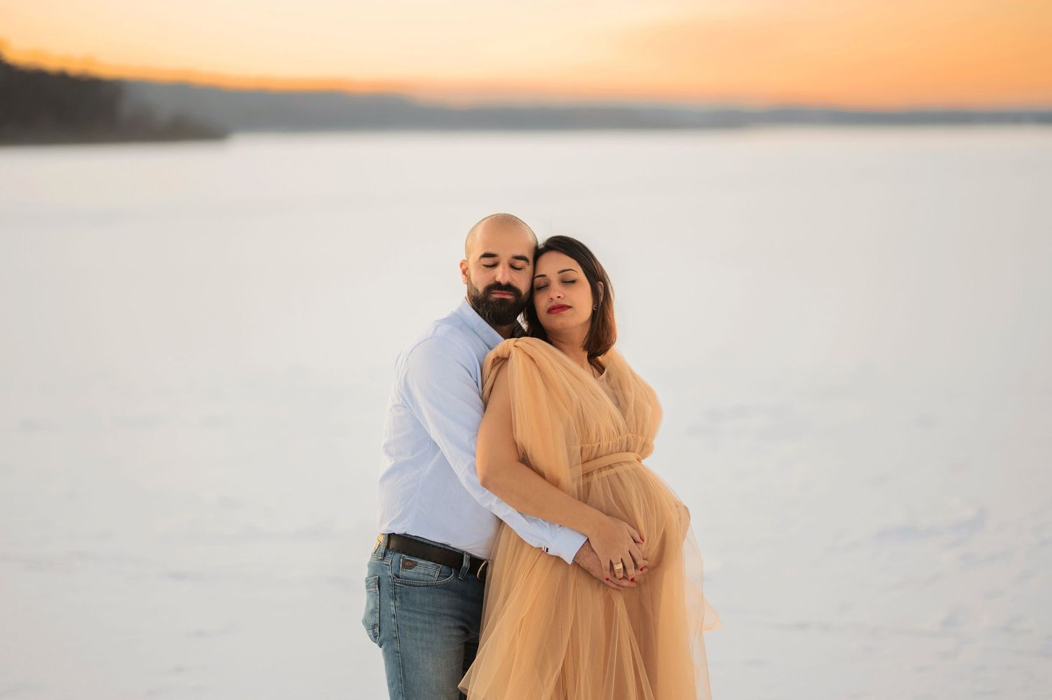 Intimate maternity portrait of a couple embracing on a snowy beach at sunset.
