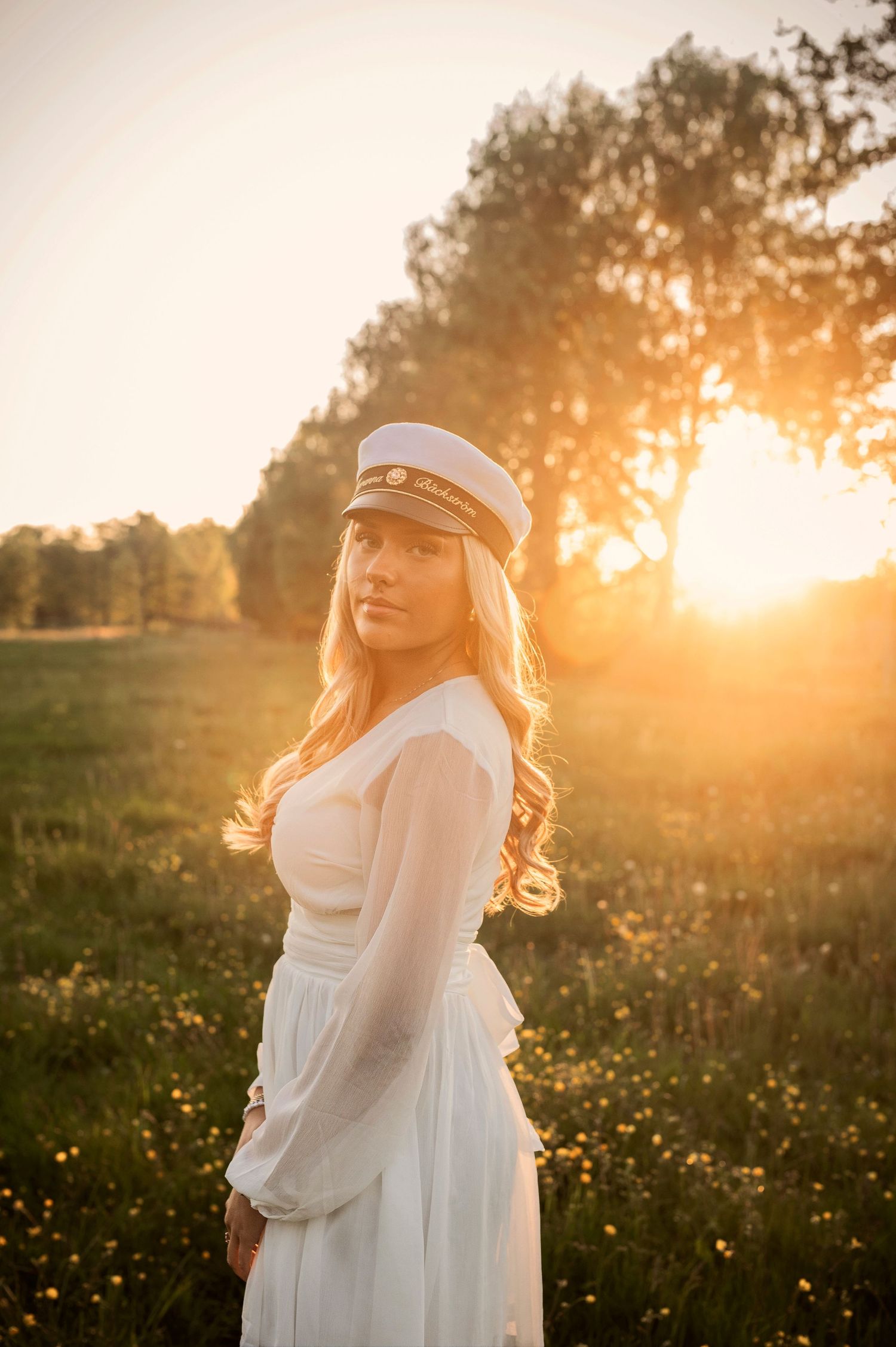Blonde woman in white dress and captain hat posing in golden sunset meadow.