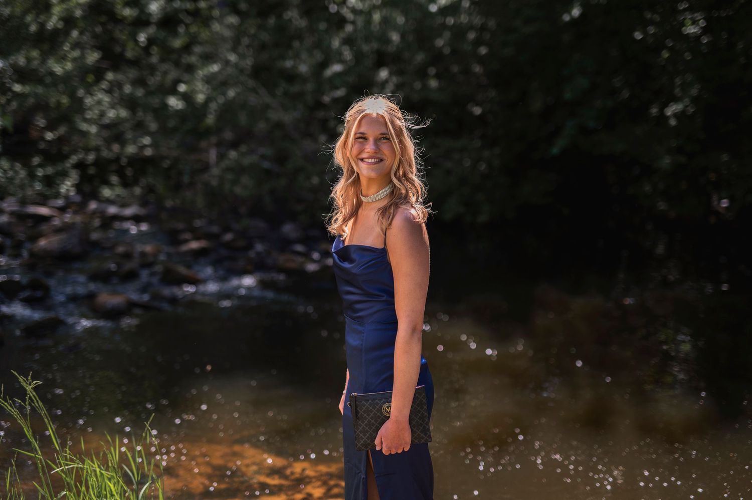A smiling blonde woman in a navy dress stands near a sunlit stream surrounded by lush green trees.