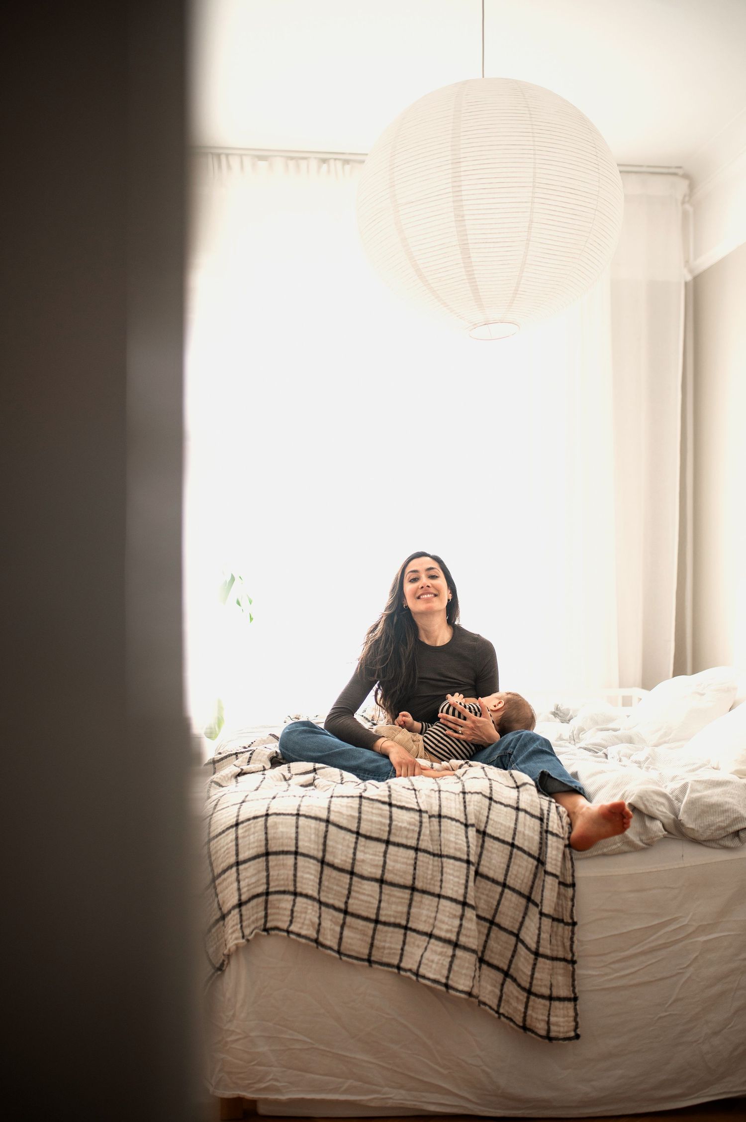 A smiling mother sits on a bed with her baby in a bright, minimalist bedroom.