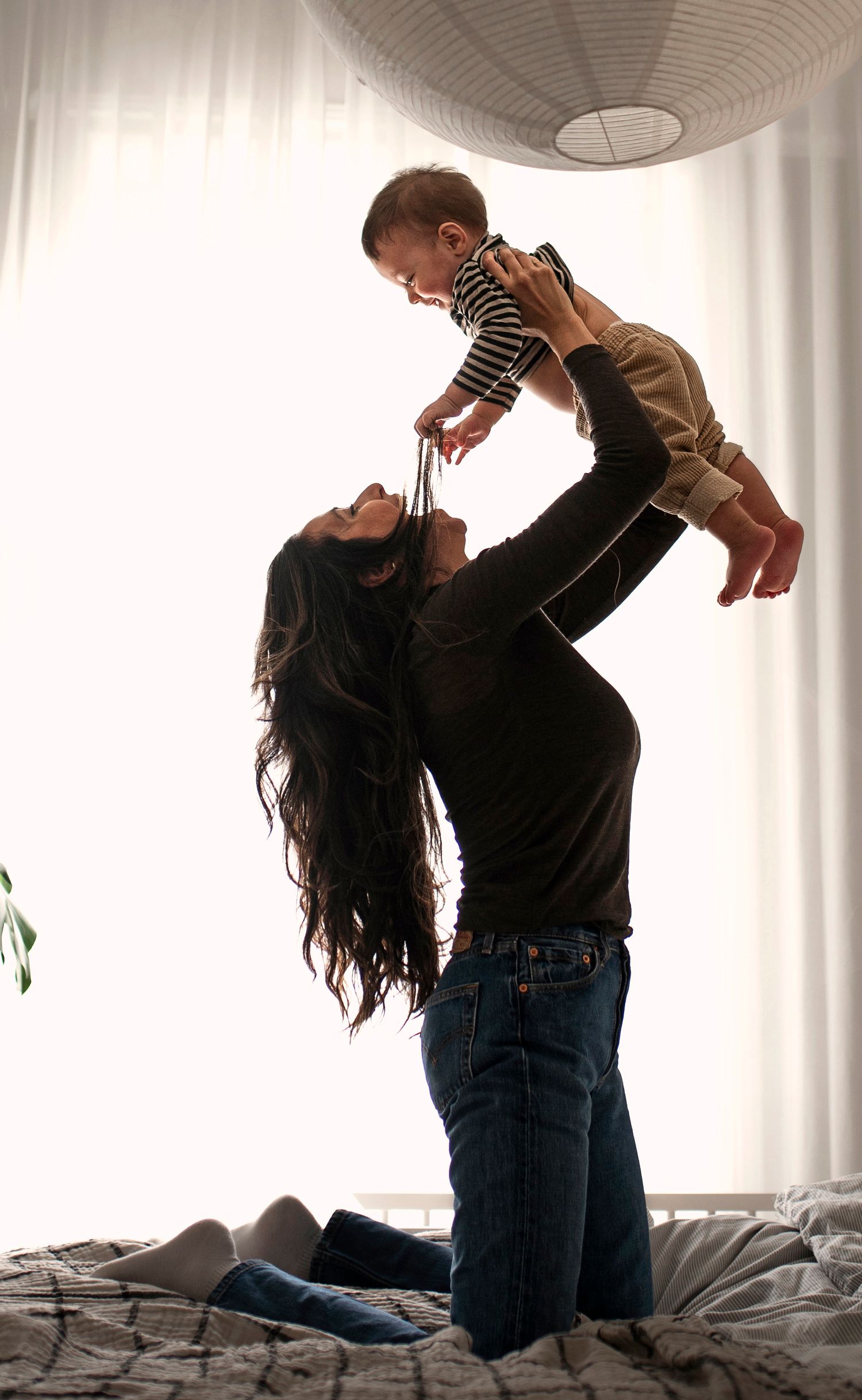 A joyful mother kneeling on a bed lifts her smiling baby overhead near a bright window.