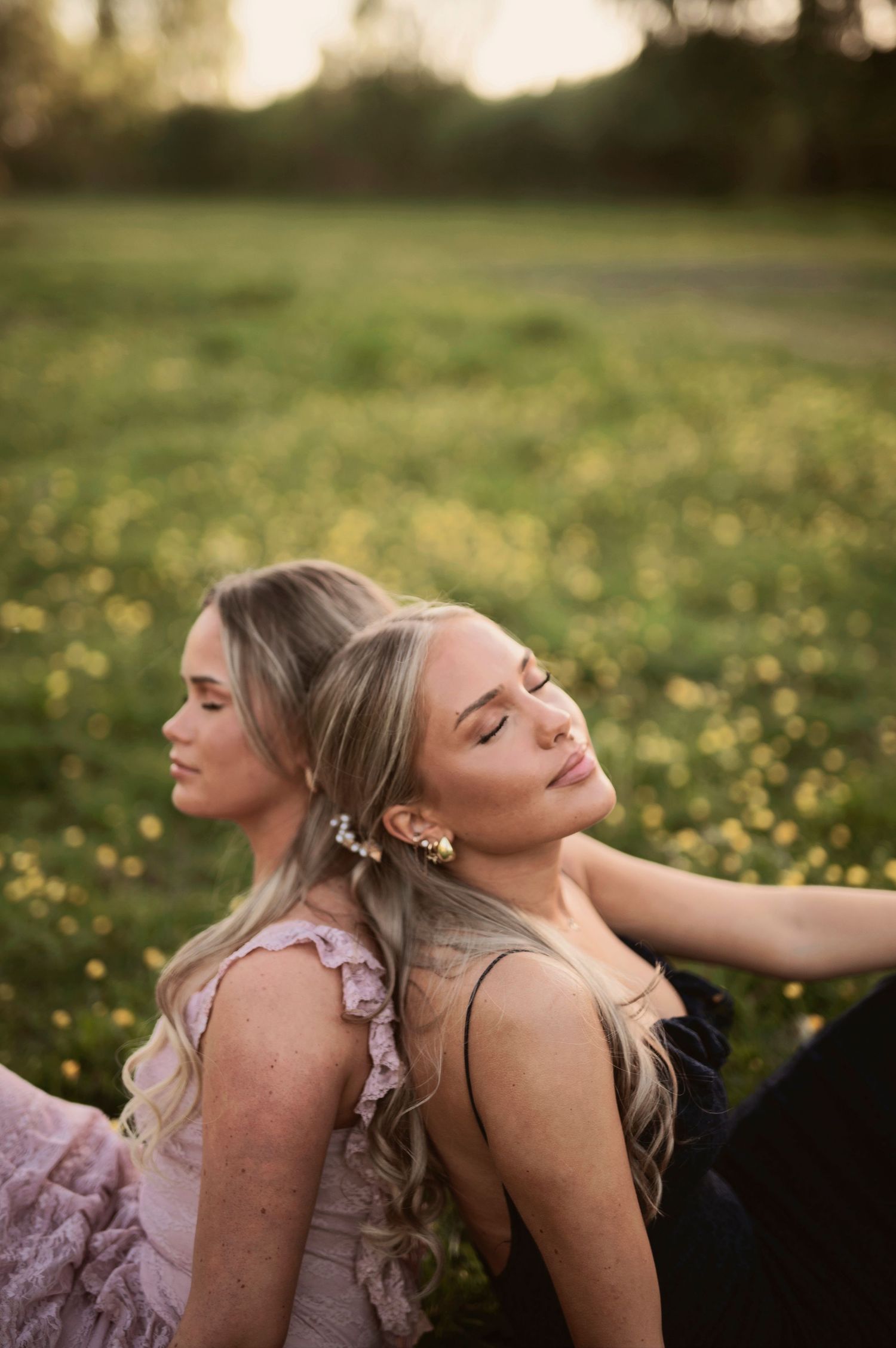Two blonde women sit back-to-back in a field of yellow wildflowers during a warm golden sunset.