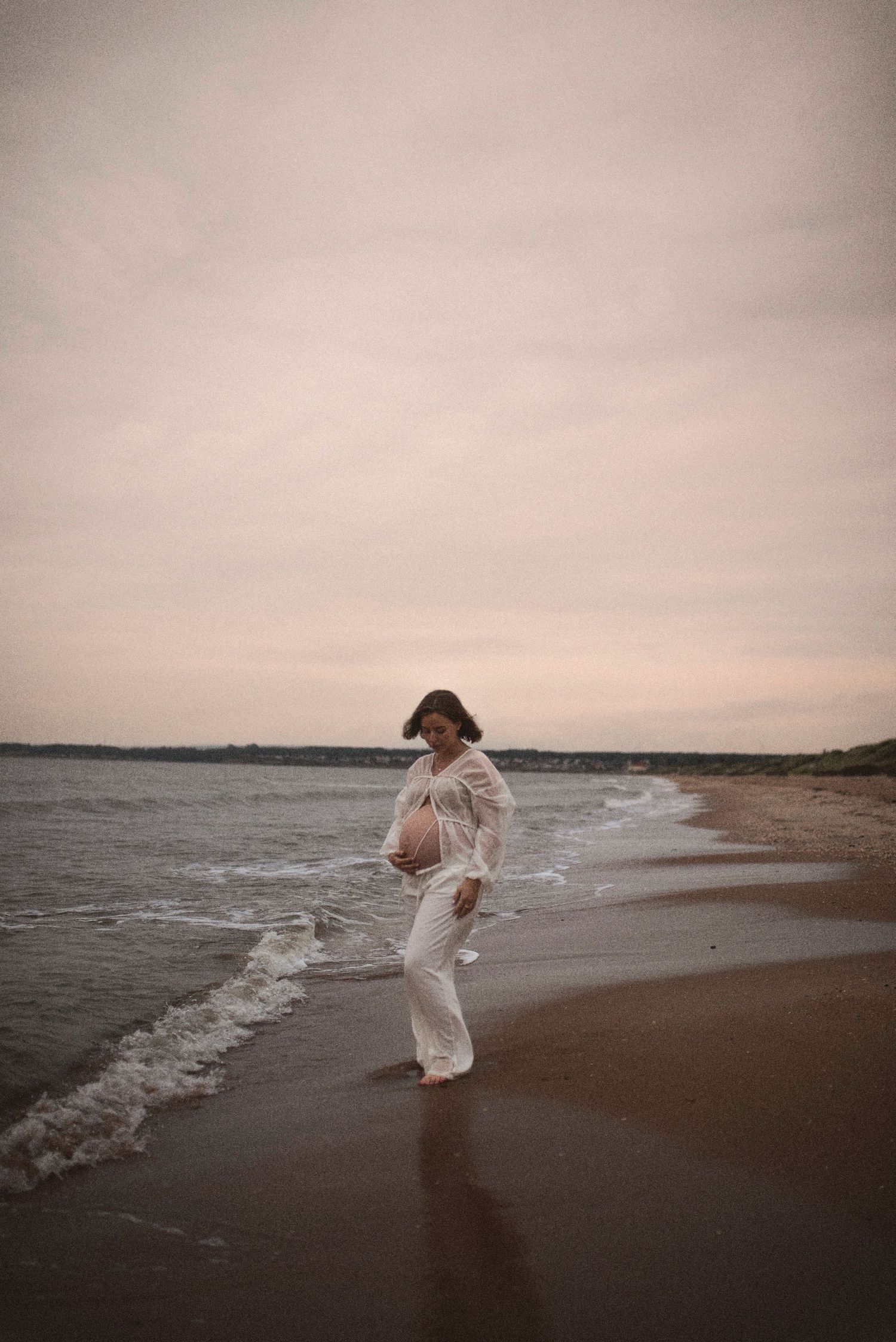 A figure in white clothing stands at the water's edge during sunset on a peaceful beach.