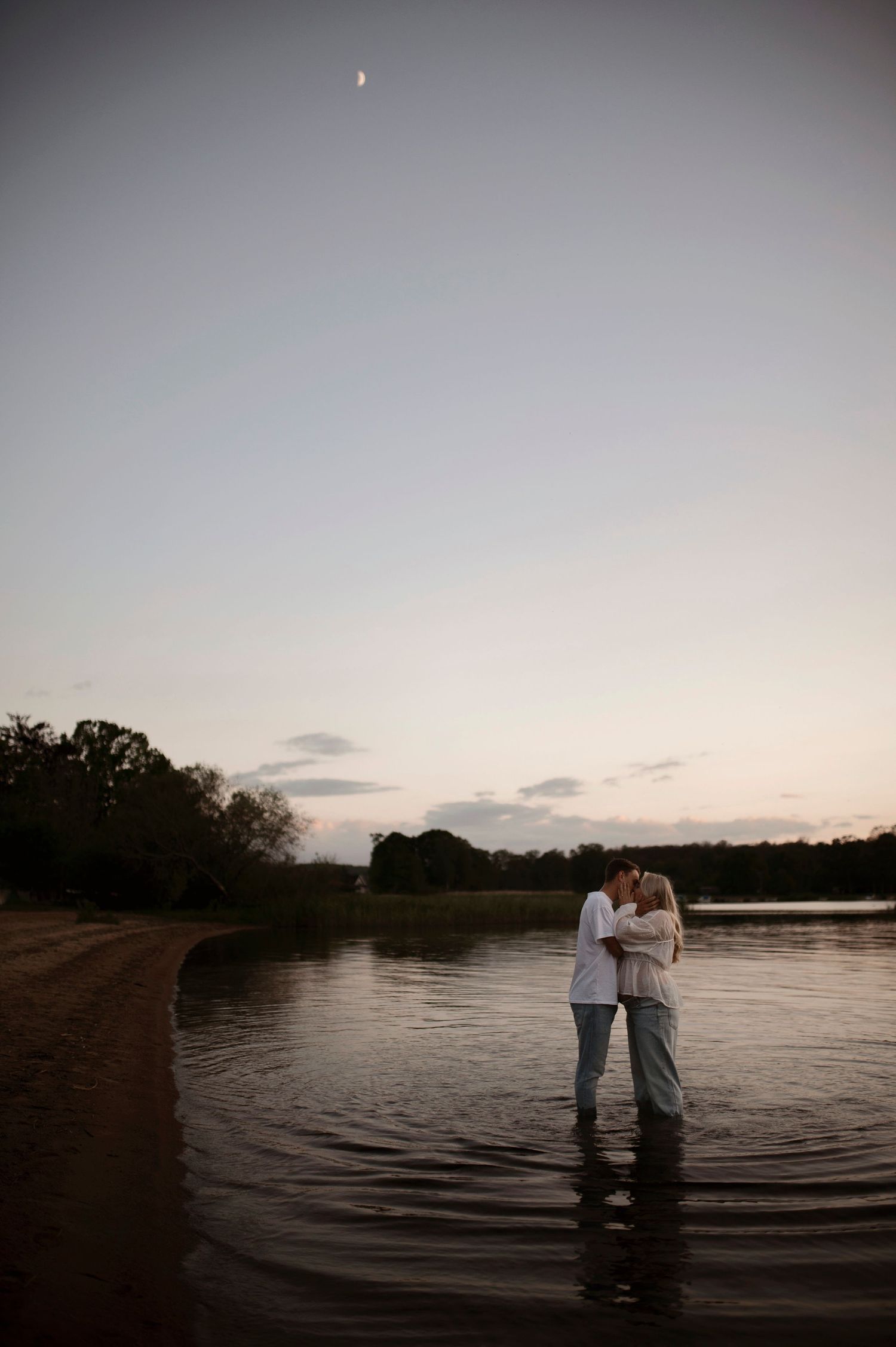 A couple embraces while standing in a lake at dusk with a crescent moon in the sky.