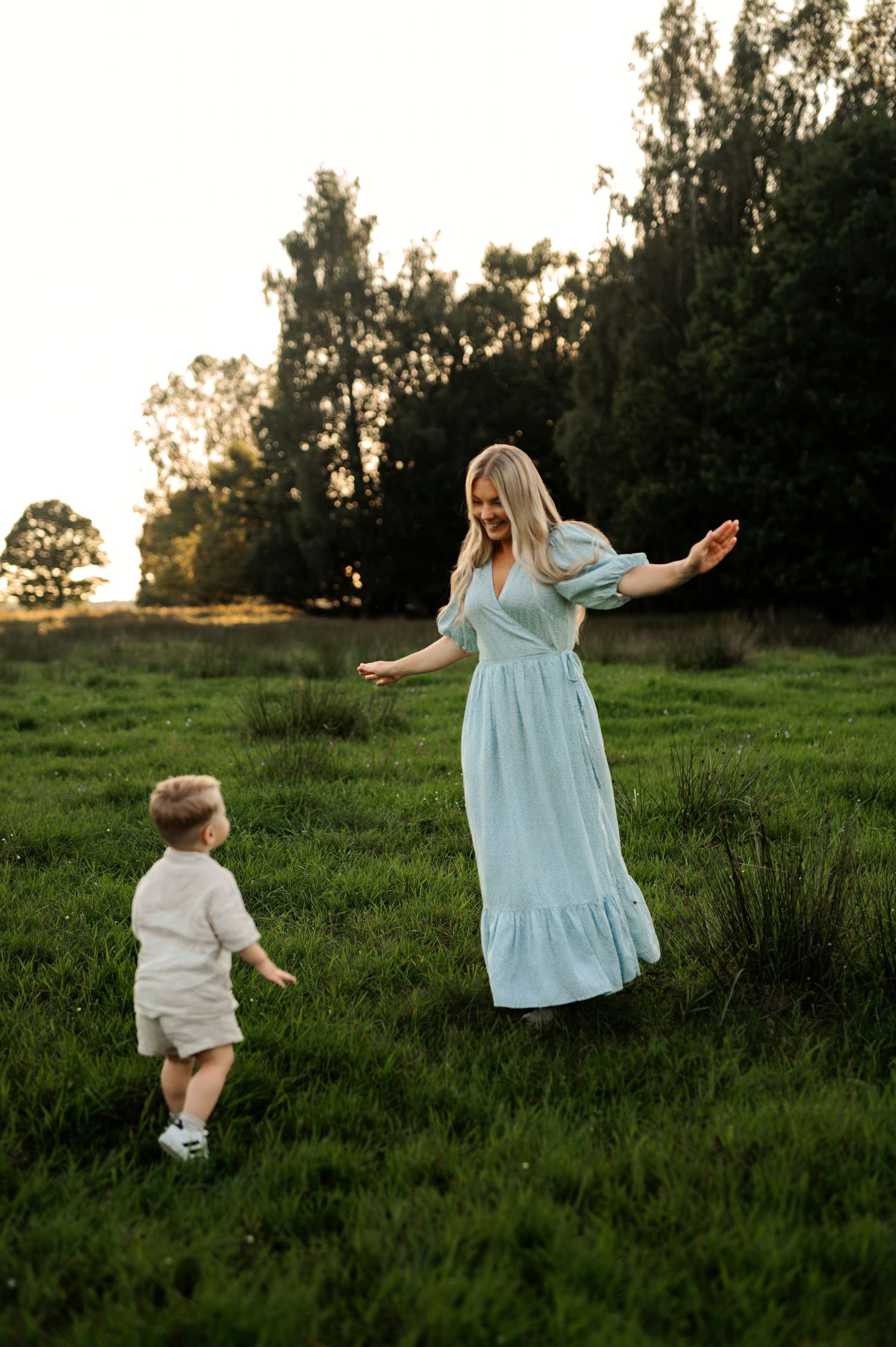 A woman in a light blue maxi dress dances with a small child in a grassy field at sunset.