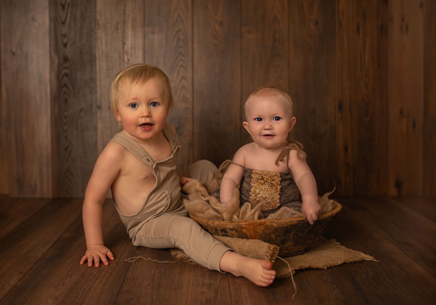 Two babies in neutral colored outfits sitting against wooden wall during portrait session.