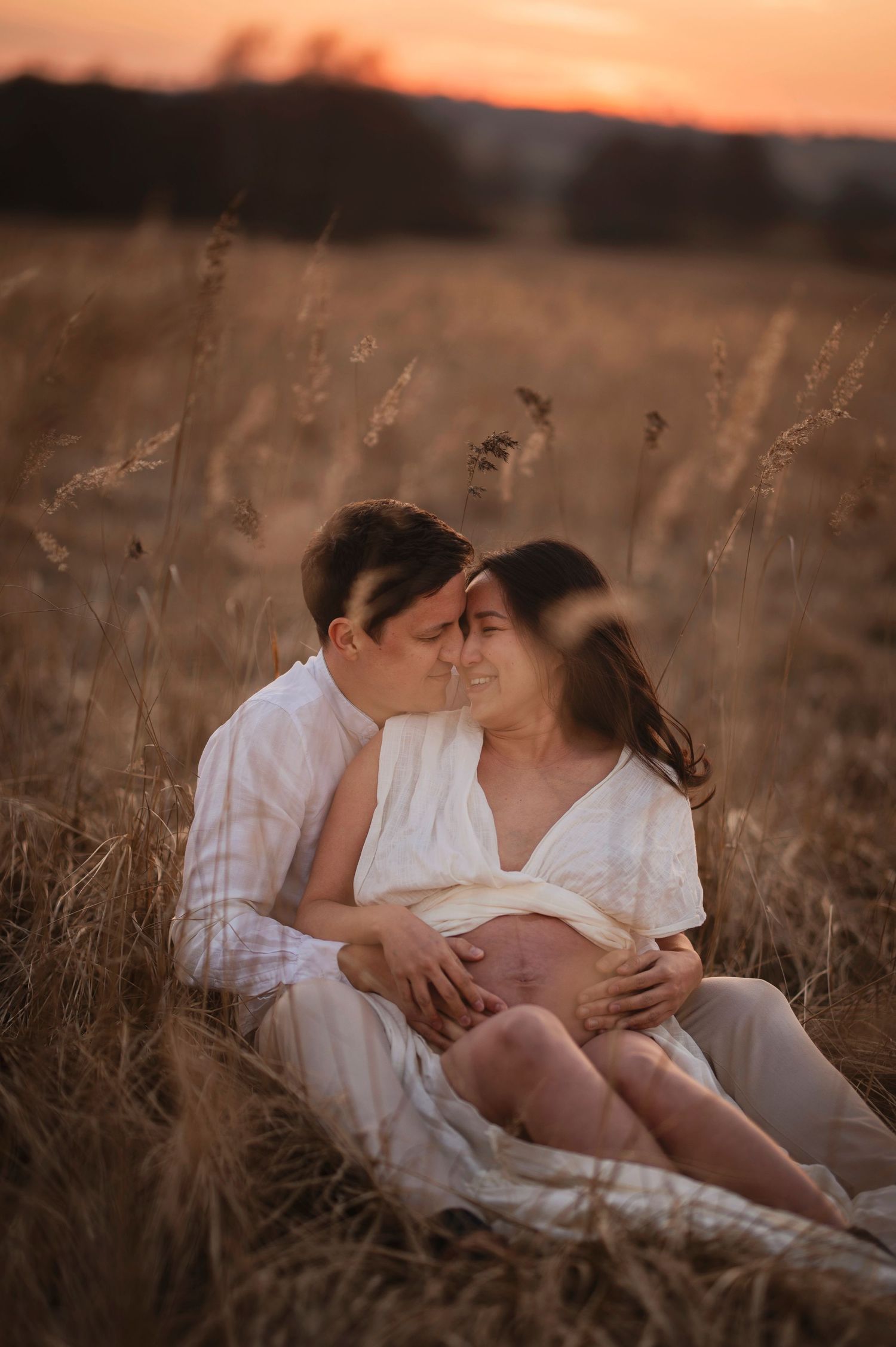 Couple in white clothing shares intimate moment while sitting in golden grass field at sunset.