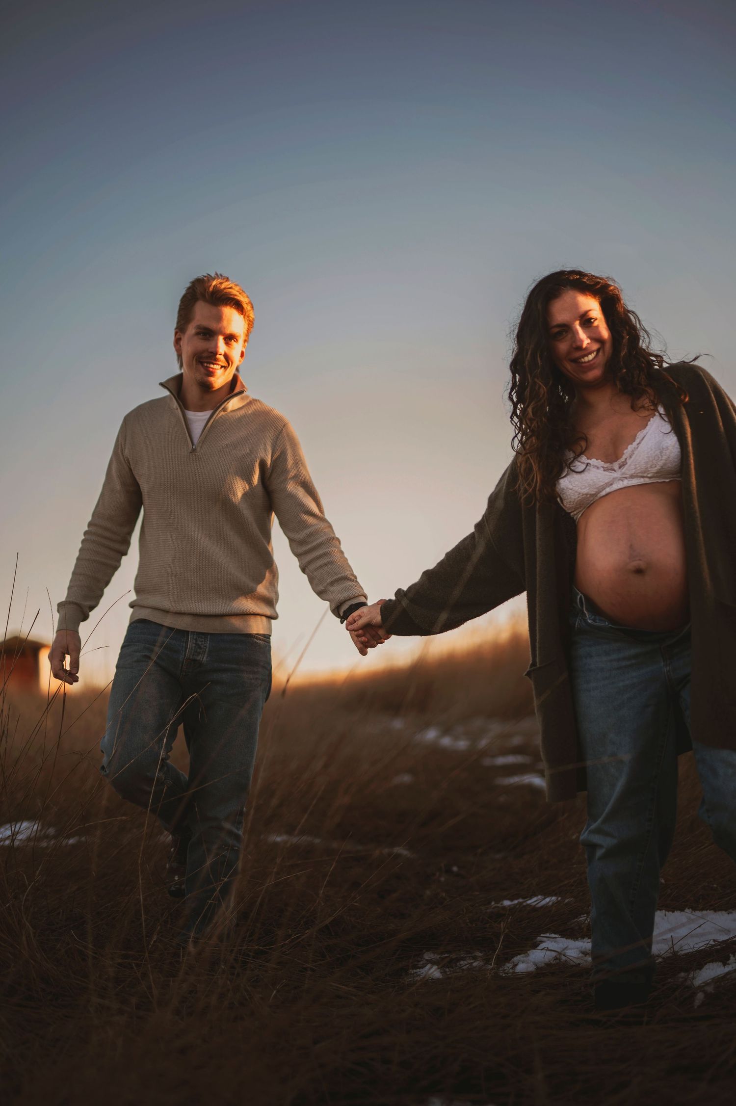 Expectant couple walking hand in hand through a field at sunset.