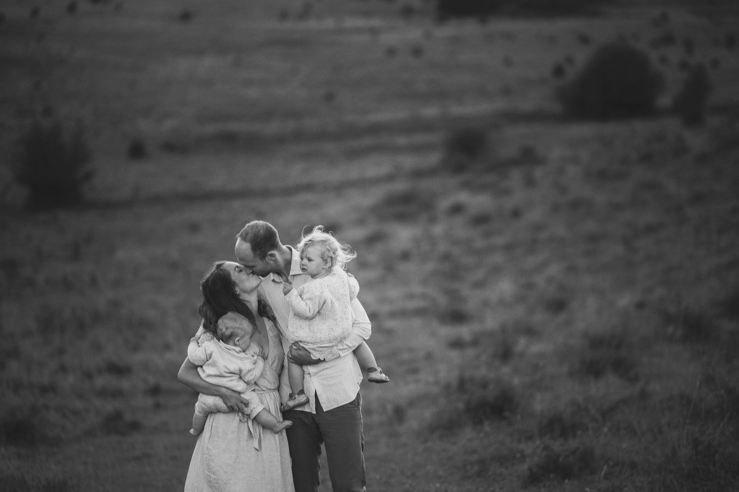 Black and white portrait of a family embracing on a hill at sunset.