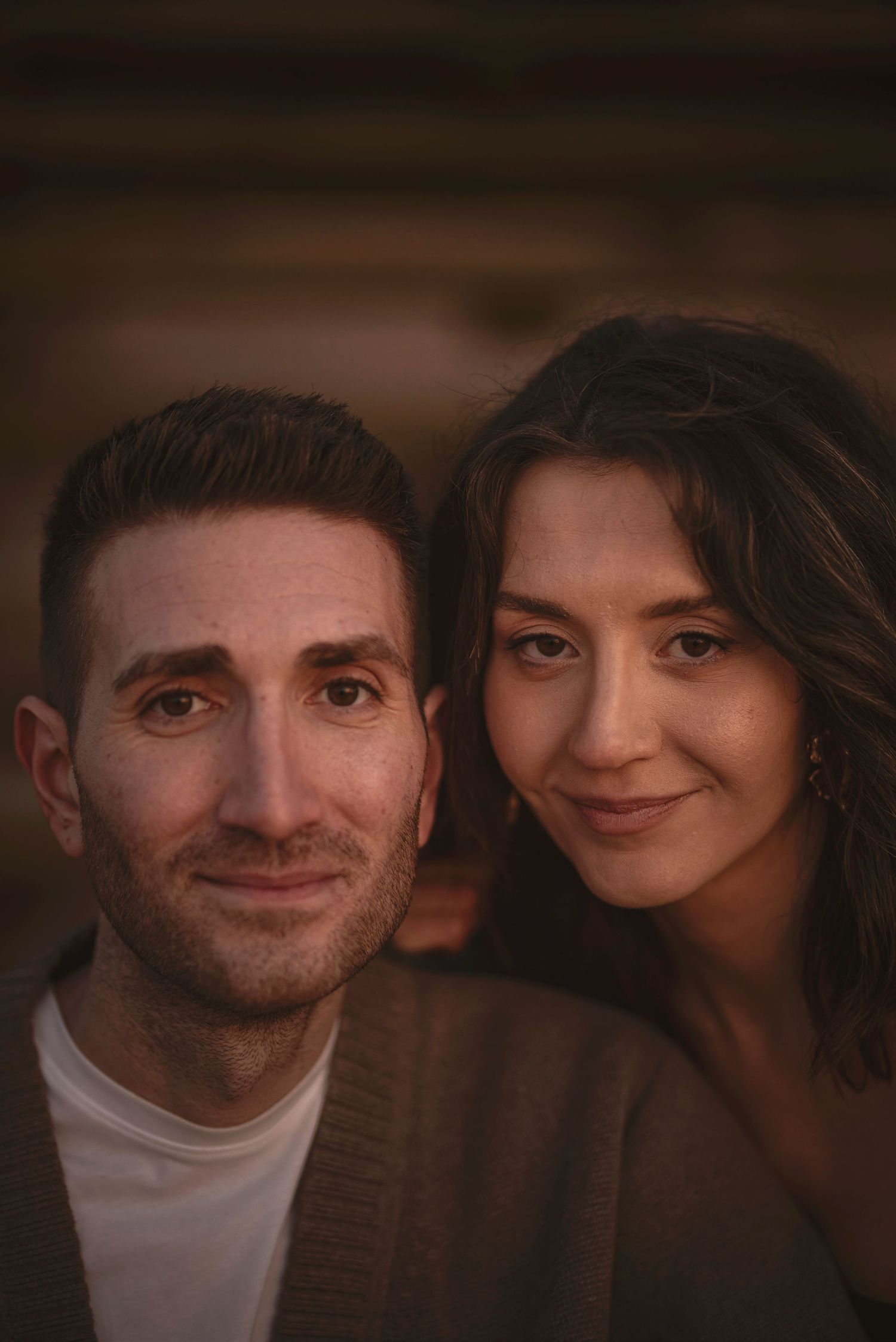 A couple shares an intimate moment in a warmly lit indoor setting with soft lighting and dark brown tones in the background.