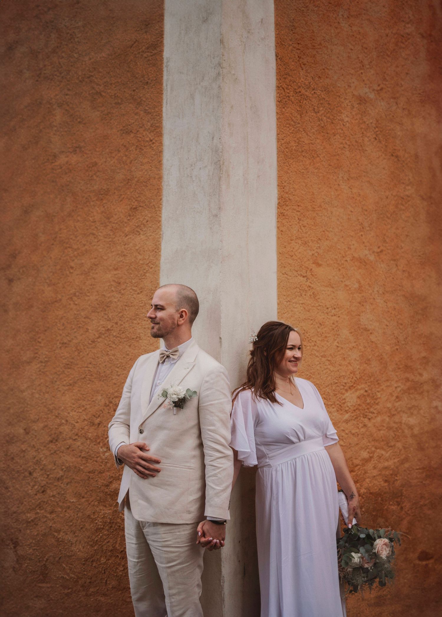 Couple in wedding attire pose against rustic orange wall holding hands and flowers.
