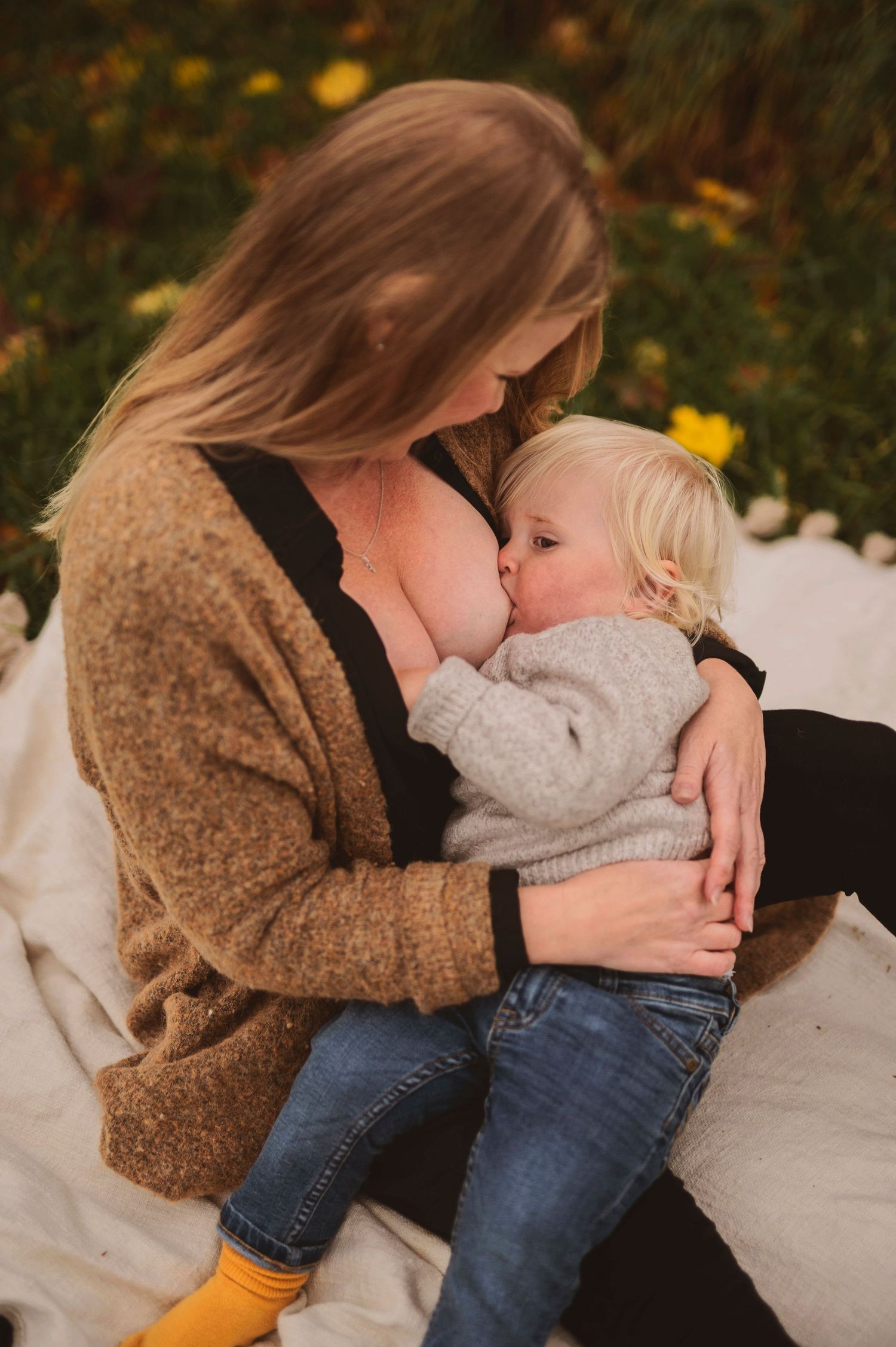 A tender moment of breastfeeding on a blanket outdoors with autumn foliage in the background.