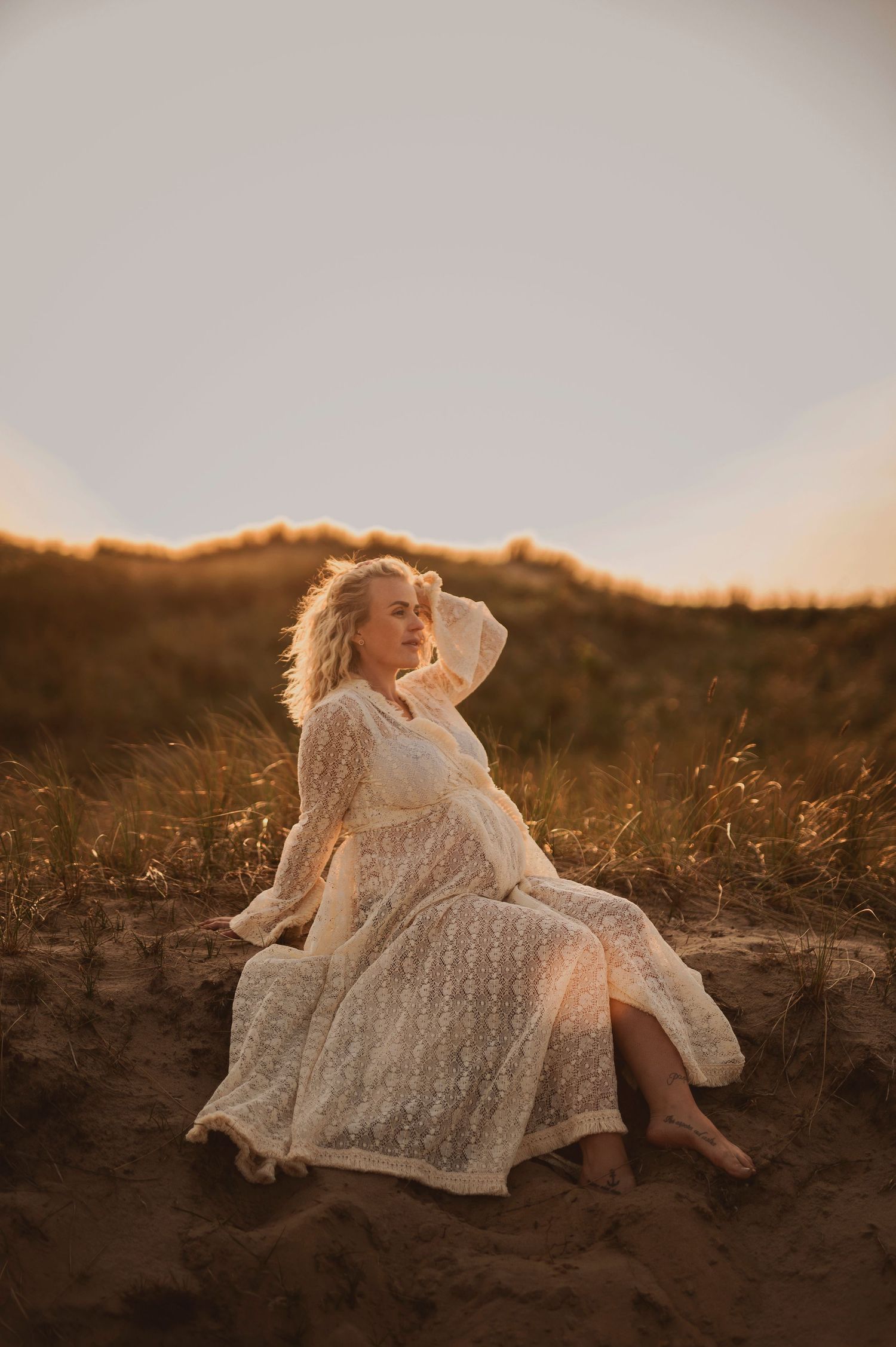 A dreamy maternity photo on sand dunes with a white lace dress at golden hour.