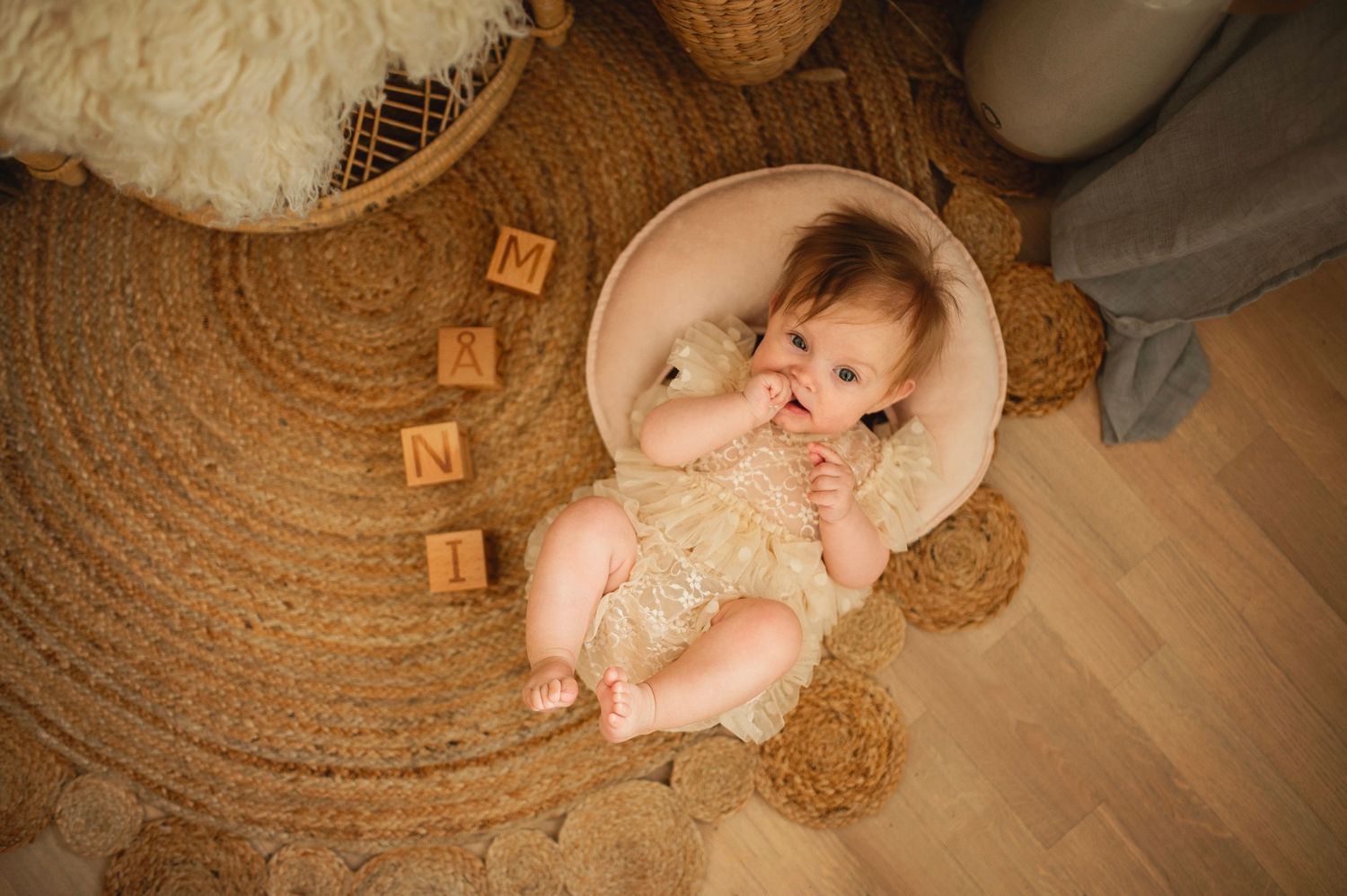 Baby lying in round basket on circular rug with wooden blocks.