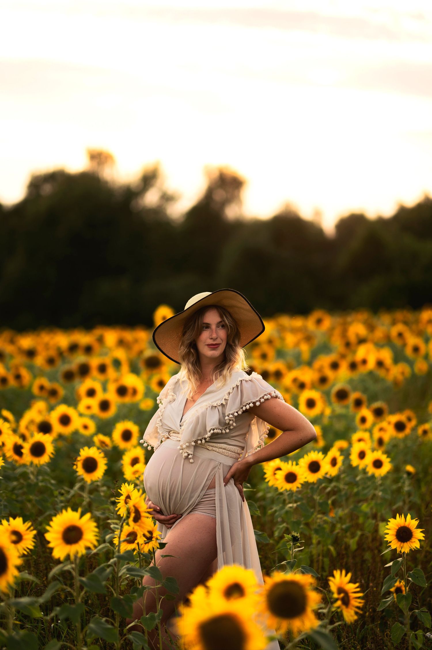 Expectant mother in wide-brimmed hat stands amid vibrant sunflowers at dusk.