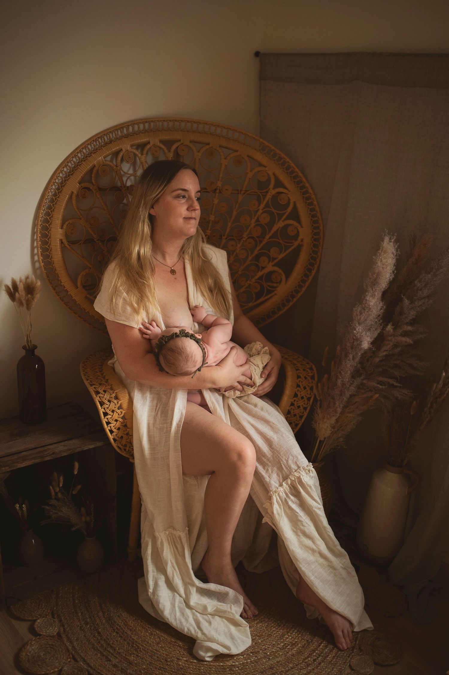 Mother in white dress sits in vintage rattan chair while nursing baby in moody natural light setting.