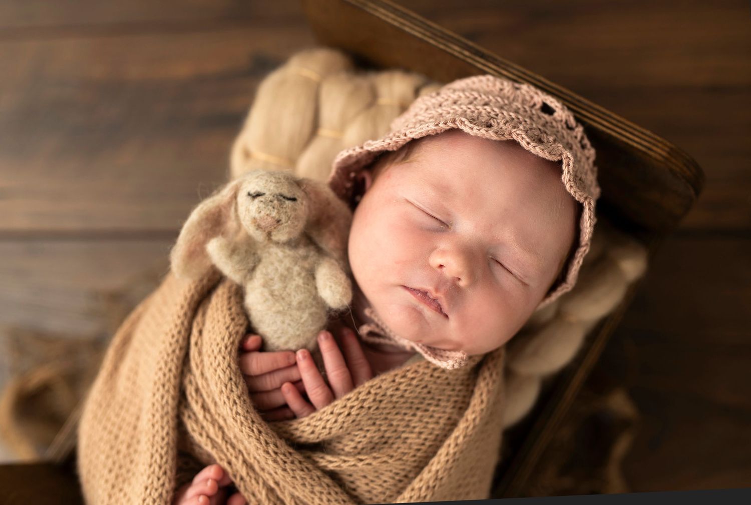 Sleeping newborn in lace bonnet cuddled with teddy bear on wooden surface.