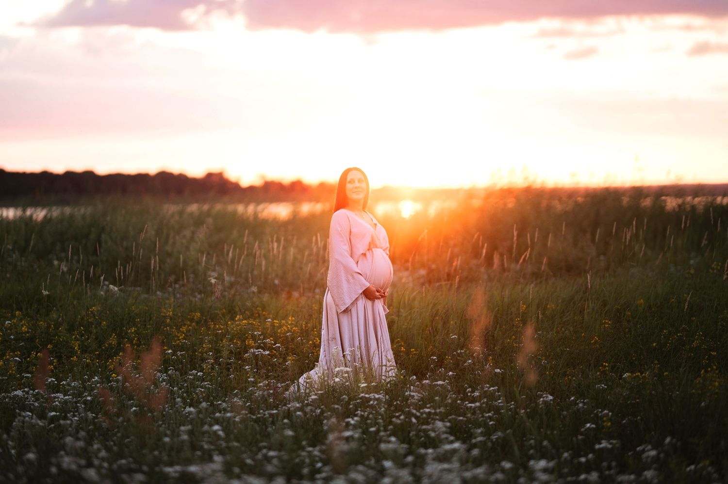 Woman in flowing dress standing in wildflower field during golden sunset.