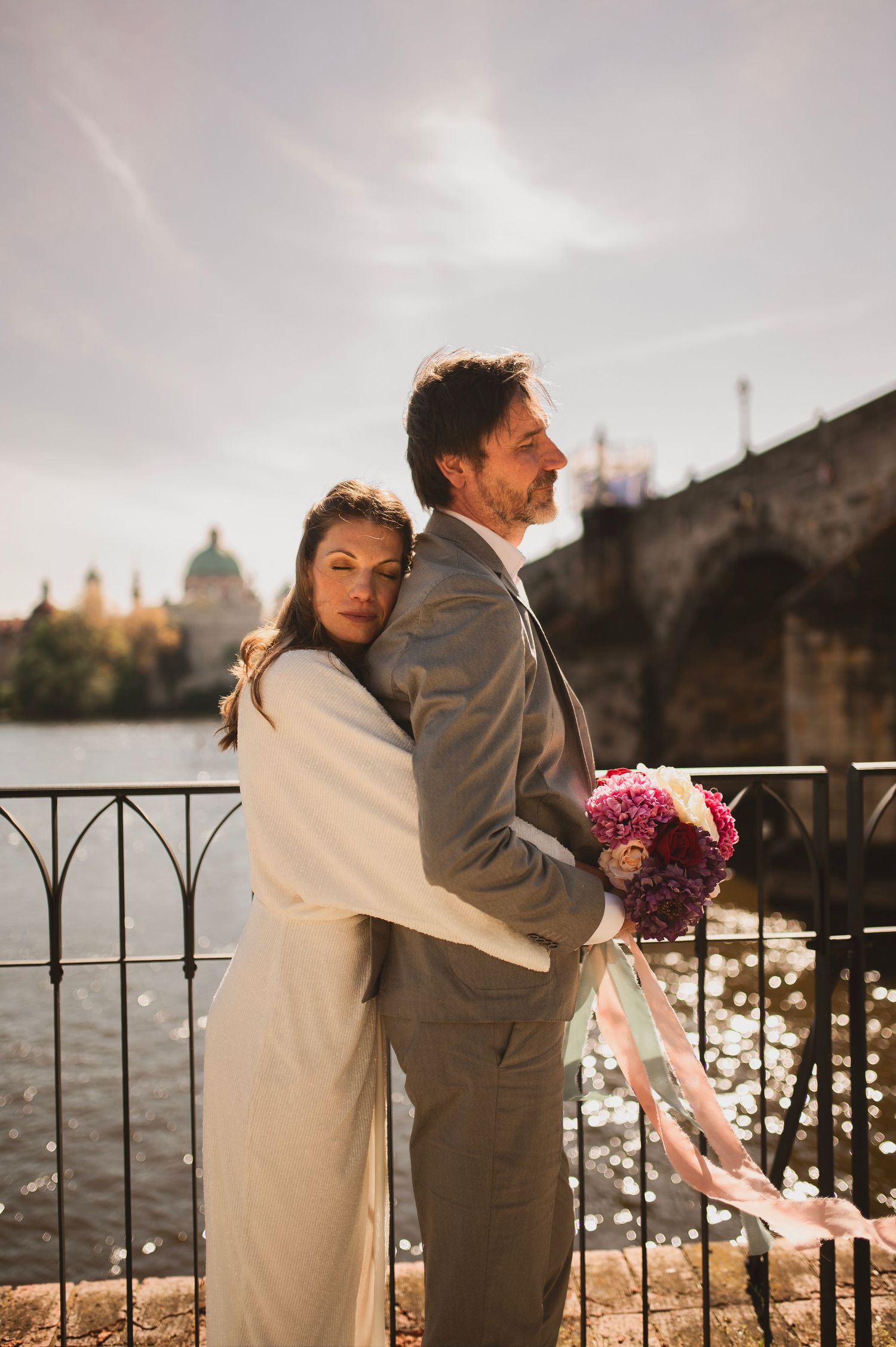Couple embraces by riverside with historic architecture and cloudy sky in background.