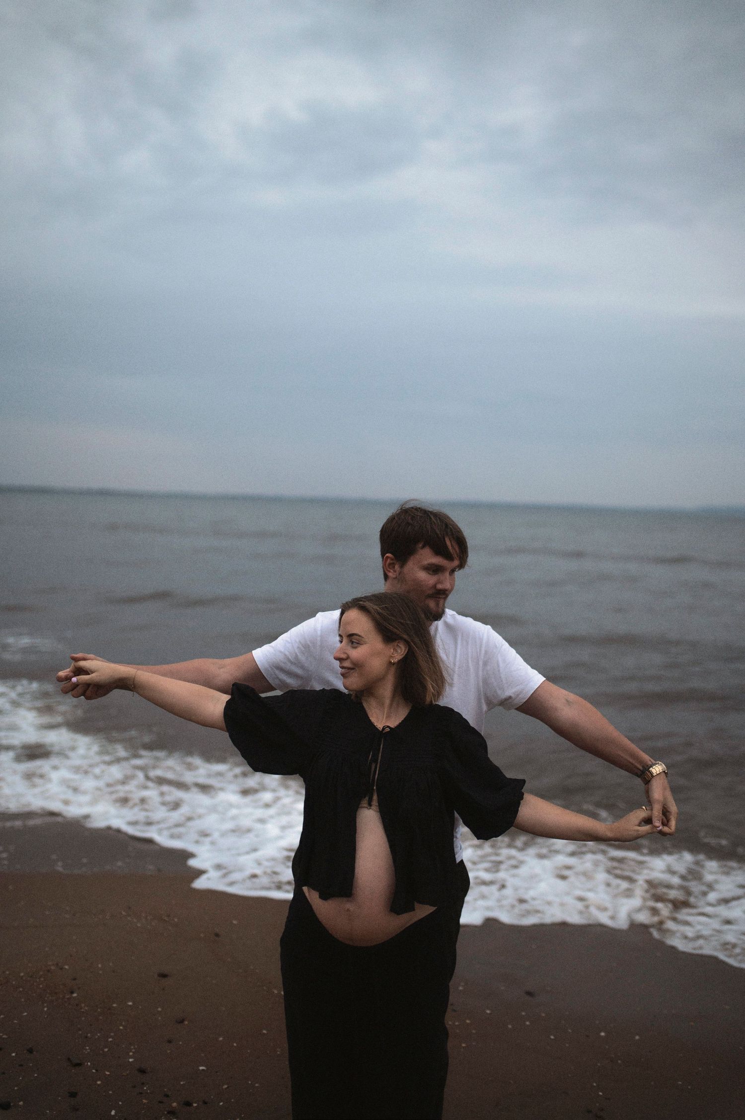 Joyful maternity moment with couple posing playfully by the ocean.