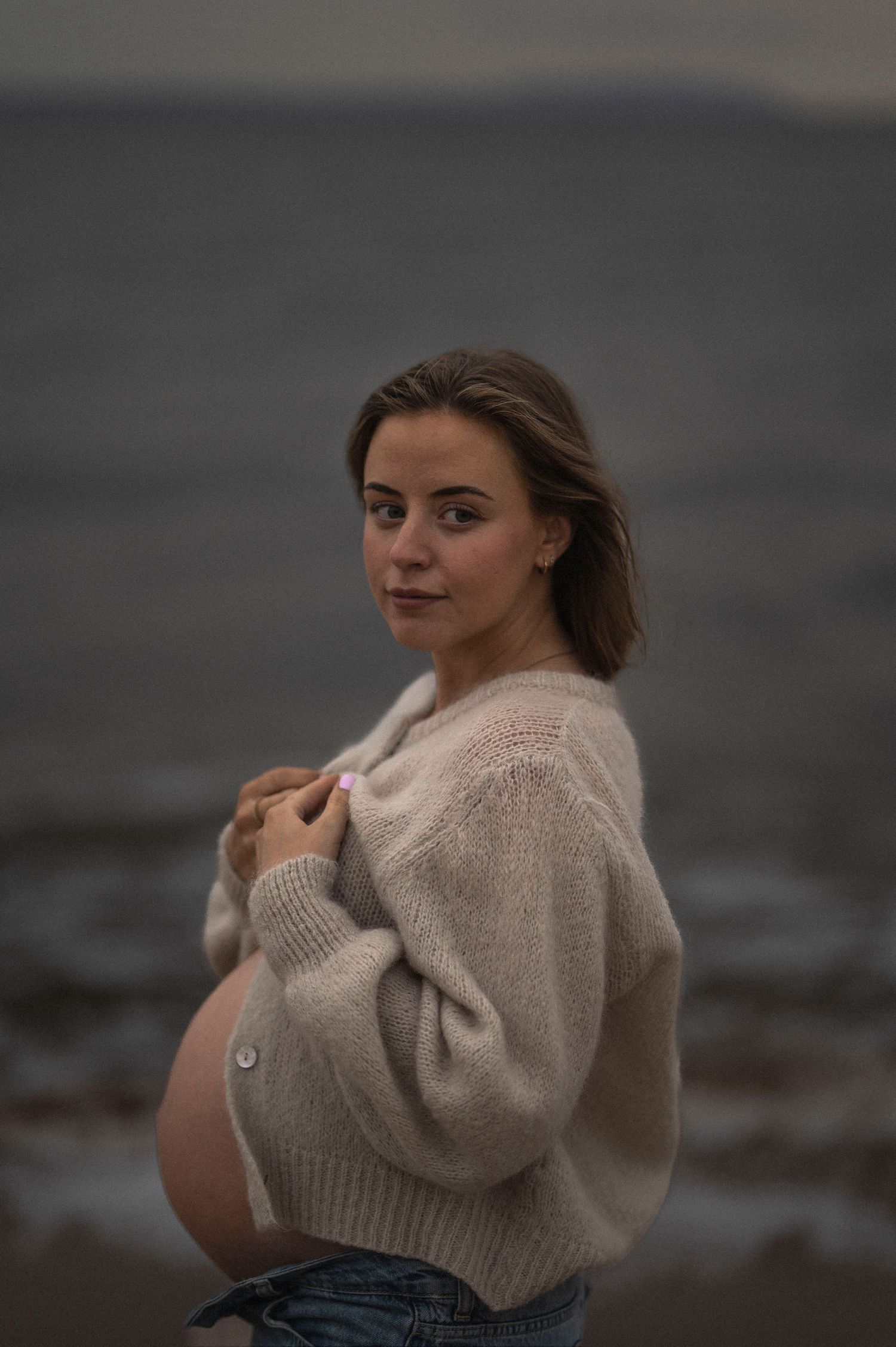 An expectant mother in a cozy sweater poses on the beach at dusk.