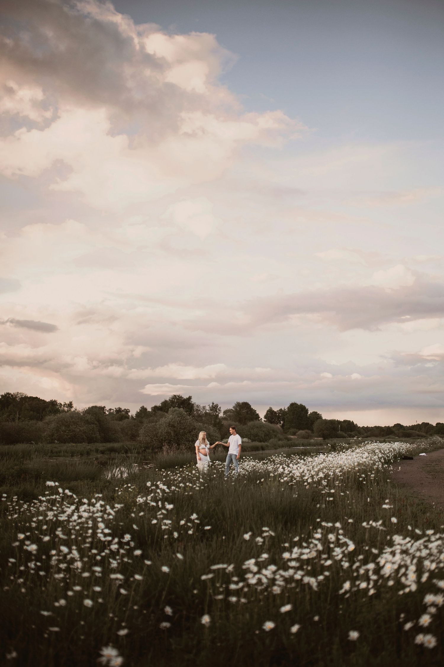 Two people stand in a dreamy meadow filled with white wildflowers at dusk under a cloudy summer sky.
