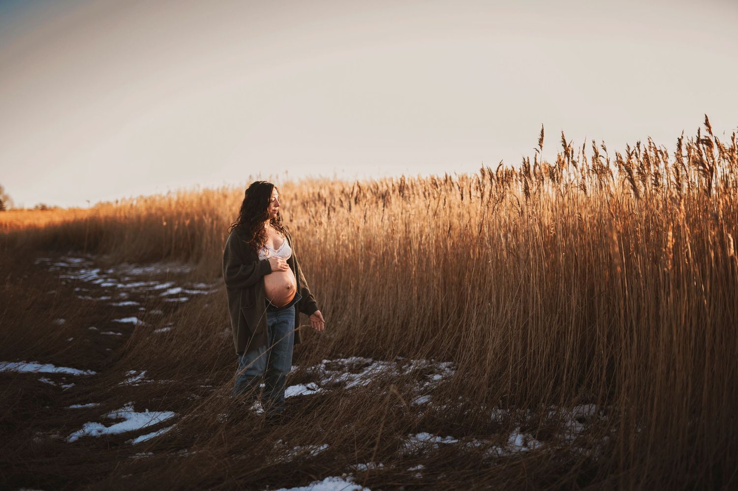 Silhouette of pregnant woman standing among tall grass at sunset with patches of snow.