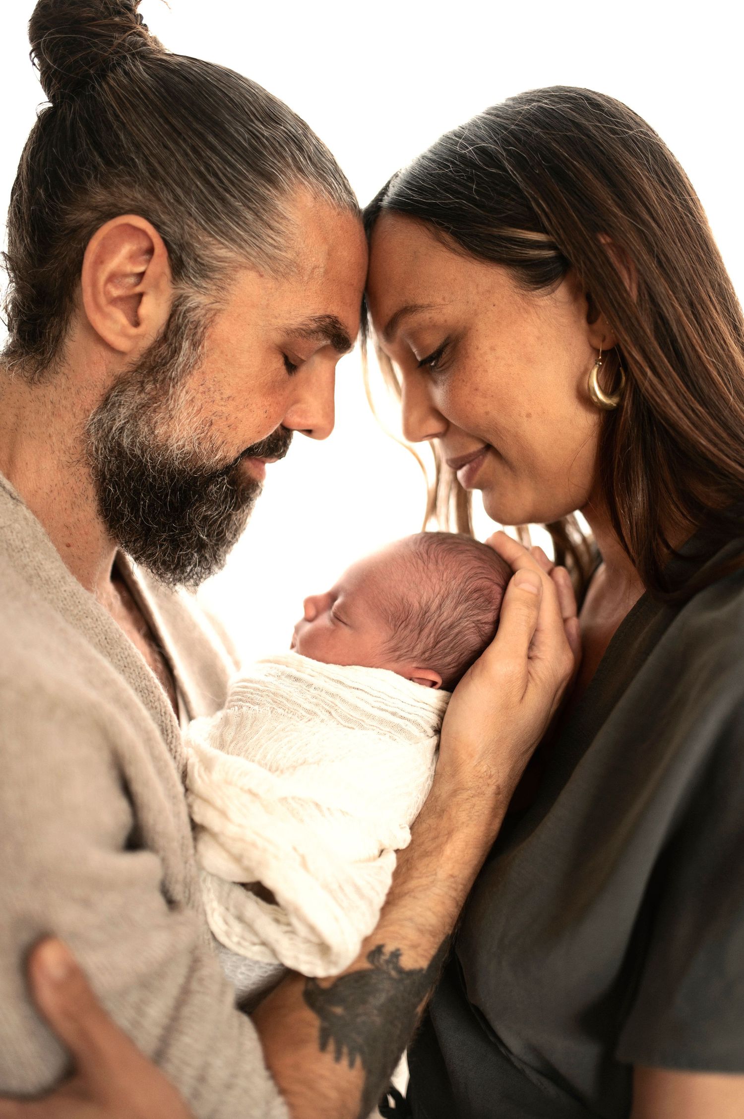 A couple tenderly holds their newborn baby, touching foreheads and smiling in a heartwarming family portrait.