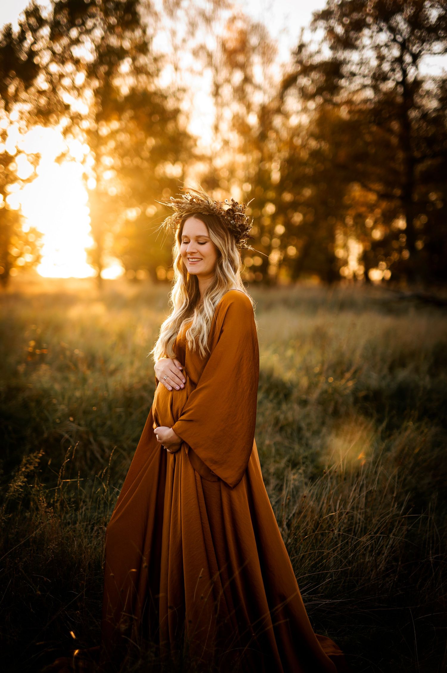 Pregnant woman in rust orange gown and floral crown stands in golden field at sunset.