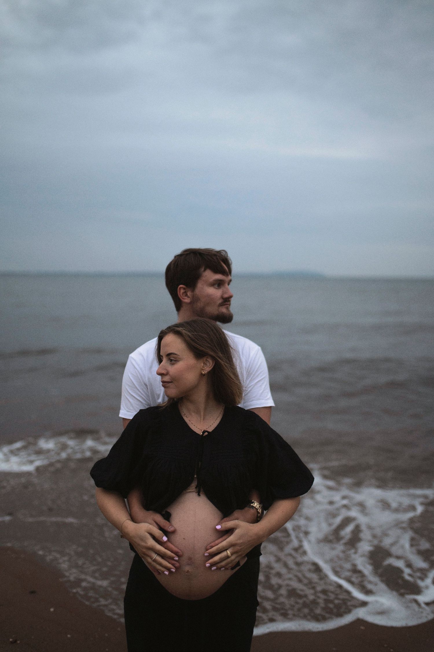 A romantic maternity photo at the beach shows an expectant couple embracing as waves lap at the shore.