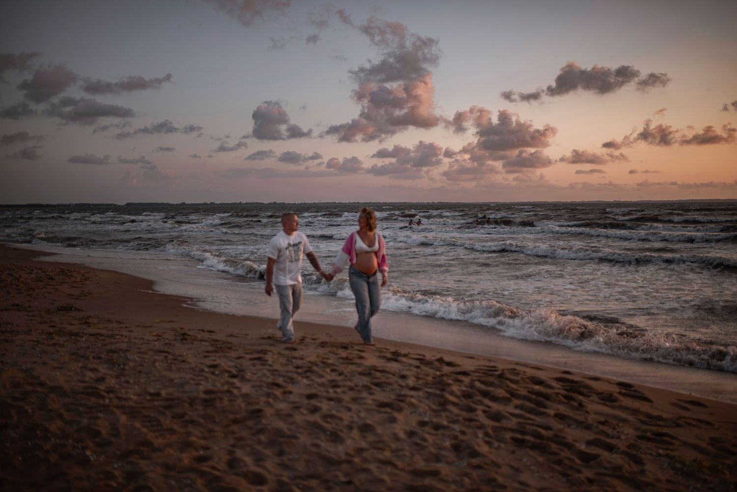 Two people hold hands while walking along a peaceful beach at sunset as waves roll onto the shore.