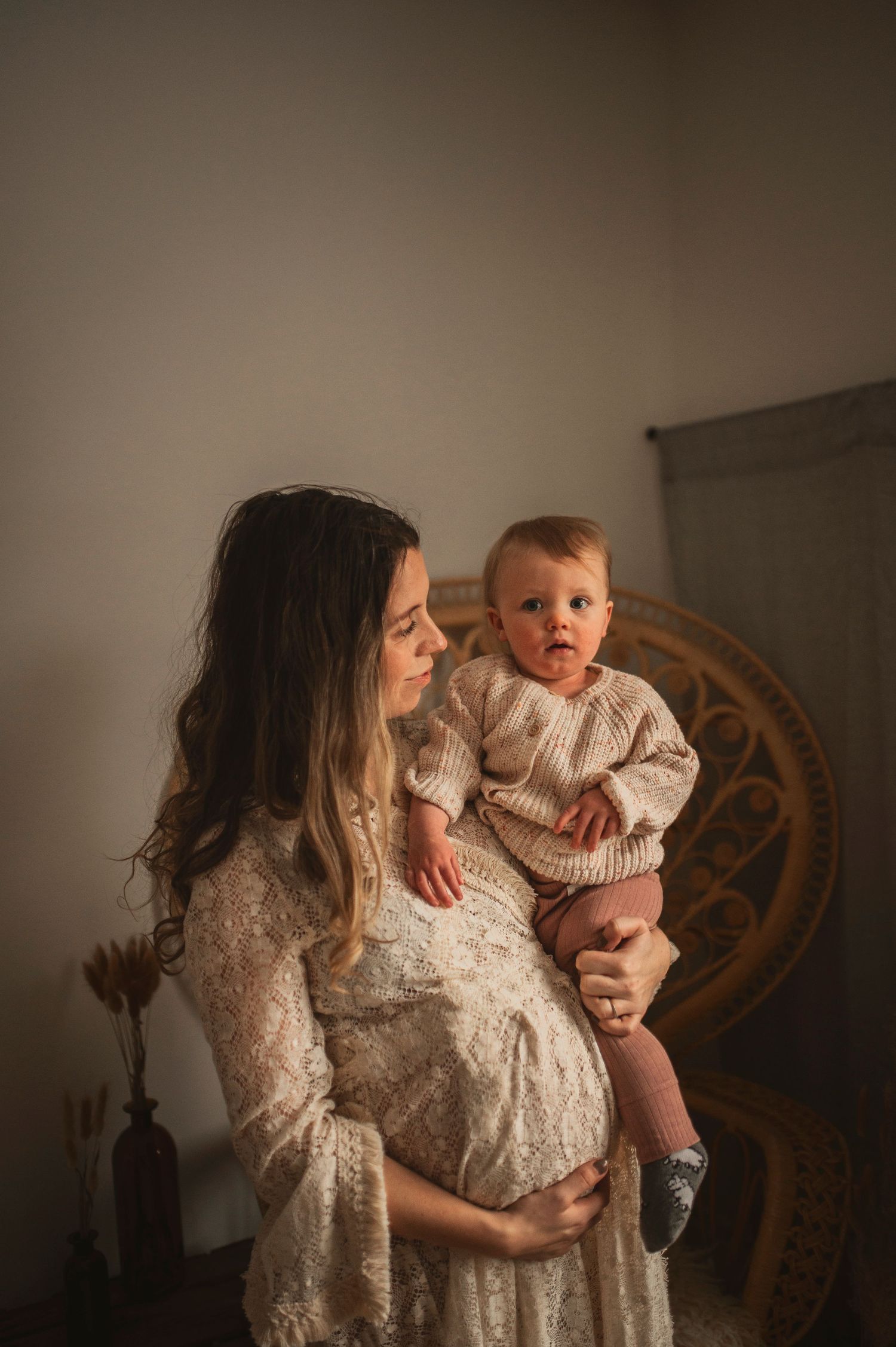 Mother in vintage lace dress holds baby in matching knit sweater beside woven chair.