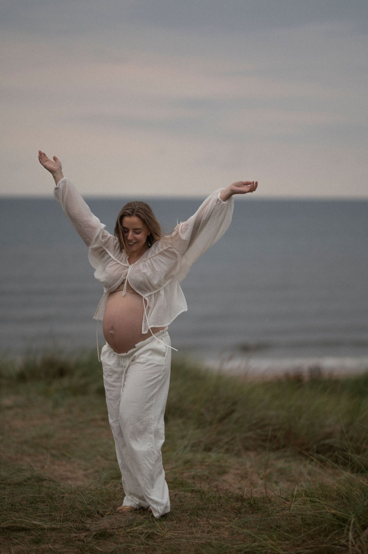 An expectant mother in flowing white attire dances joyfully on a coastal cliff at dusk.