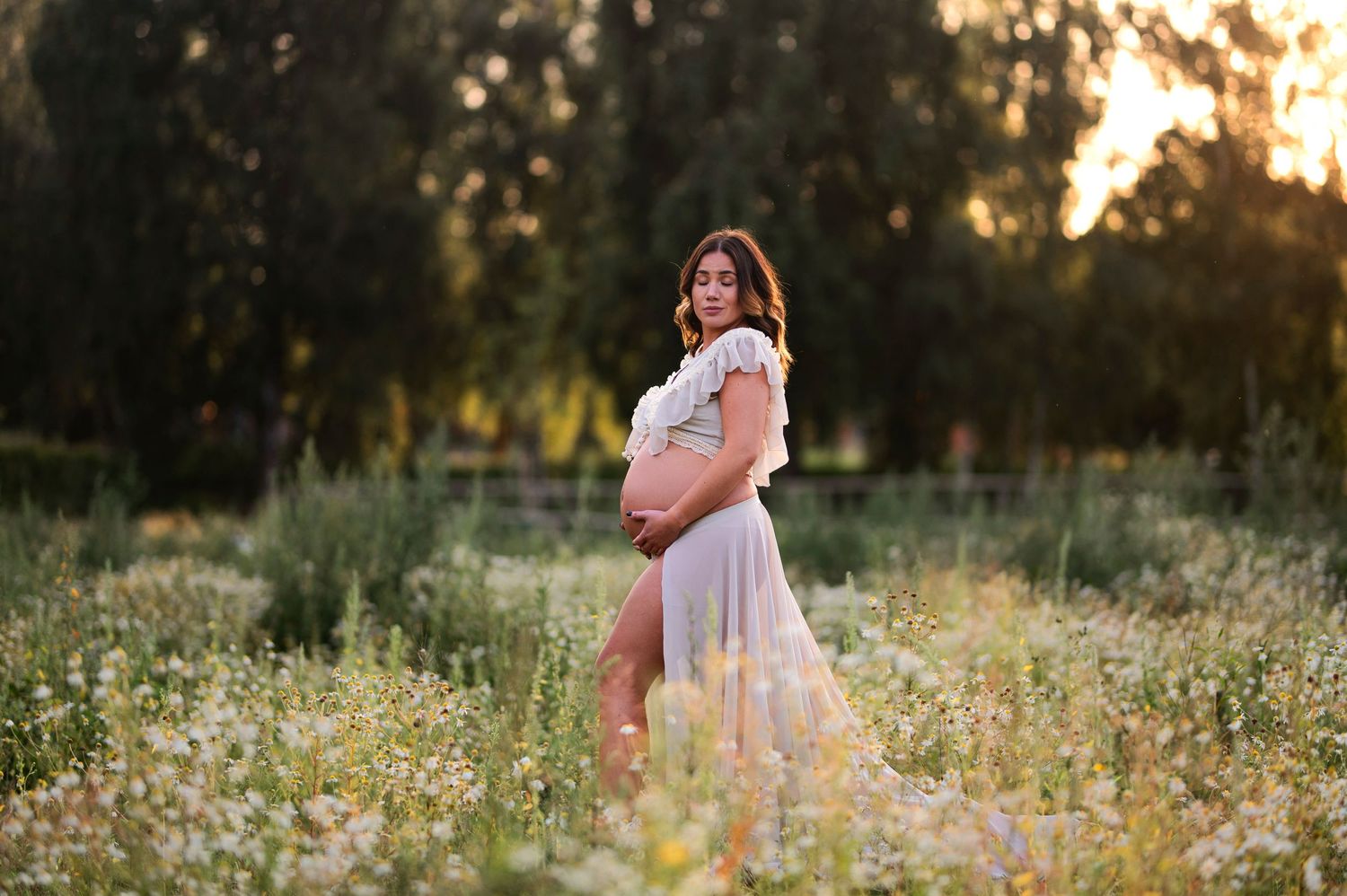 Ethereal maternity portrait in white dress amid wildflower meadow at golden hour.