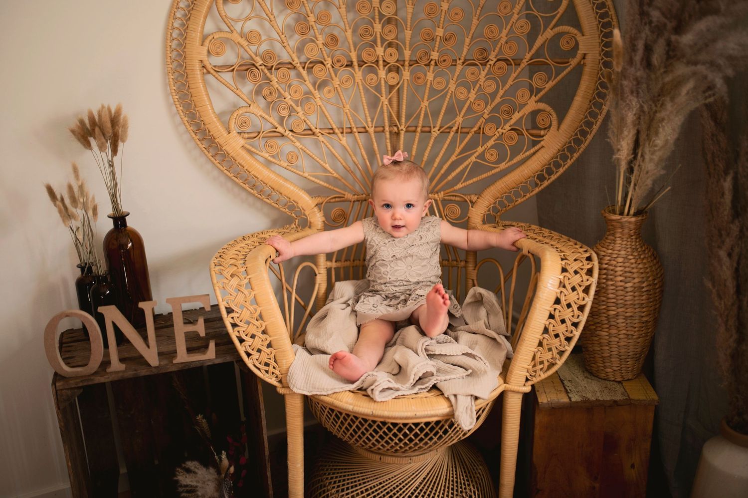 A baby sits in a vintage peacock chair surrounded by boho decor and a wooden ONE sign.