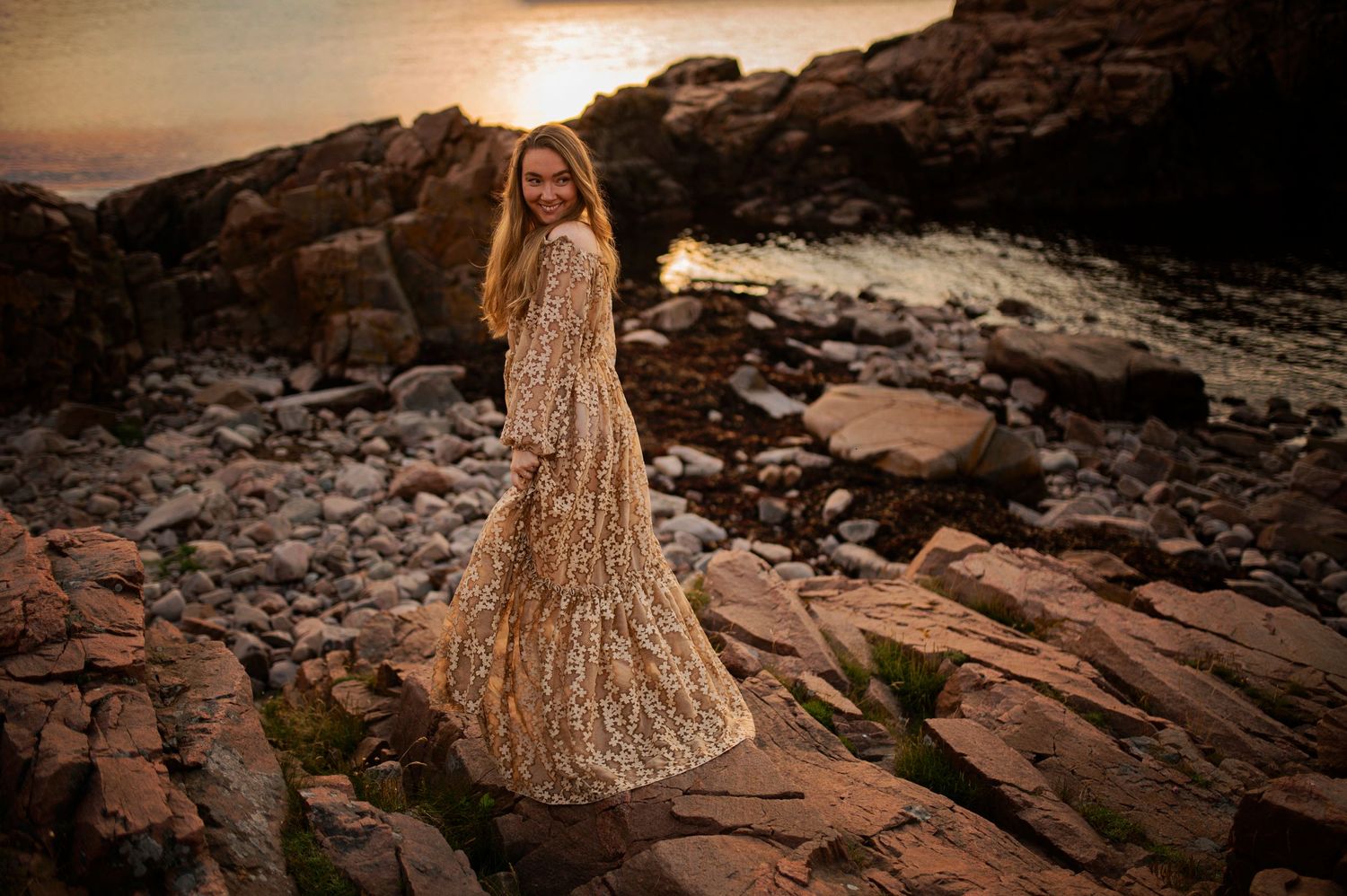 Person in sparkling gold dress posing on rocky coastline at sunset.