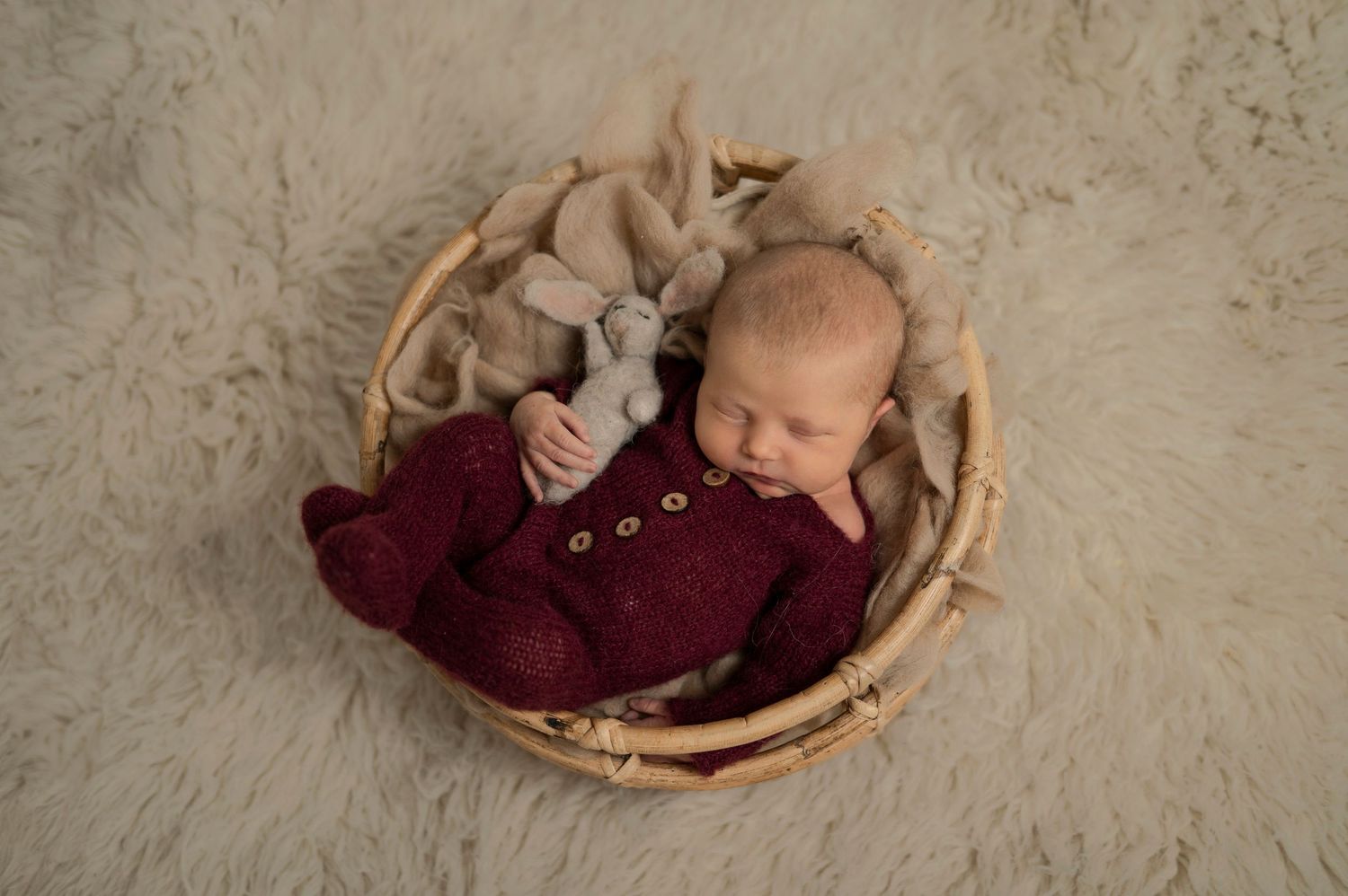Newborn baby in burgundy knit outfit sleeping in wicker basket on fur rug.
