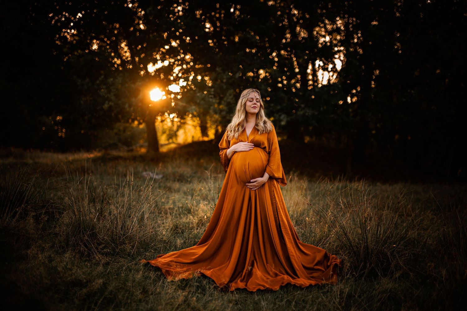 Ethereal maternity photoshoot in a rust-colored gown against golden sunset in outdoor field setting.