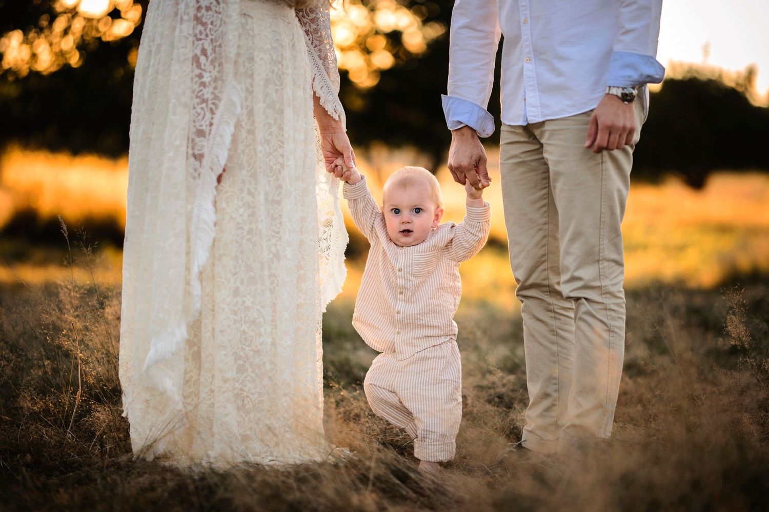 A baby takes first steps while holding parents' hands in a sunlit meadow.